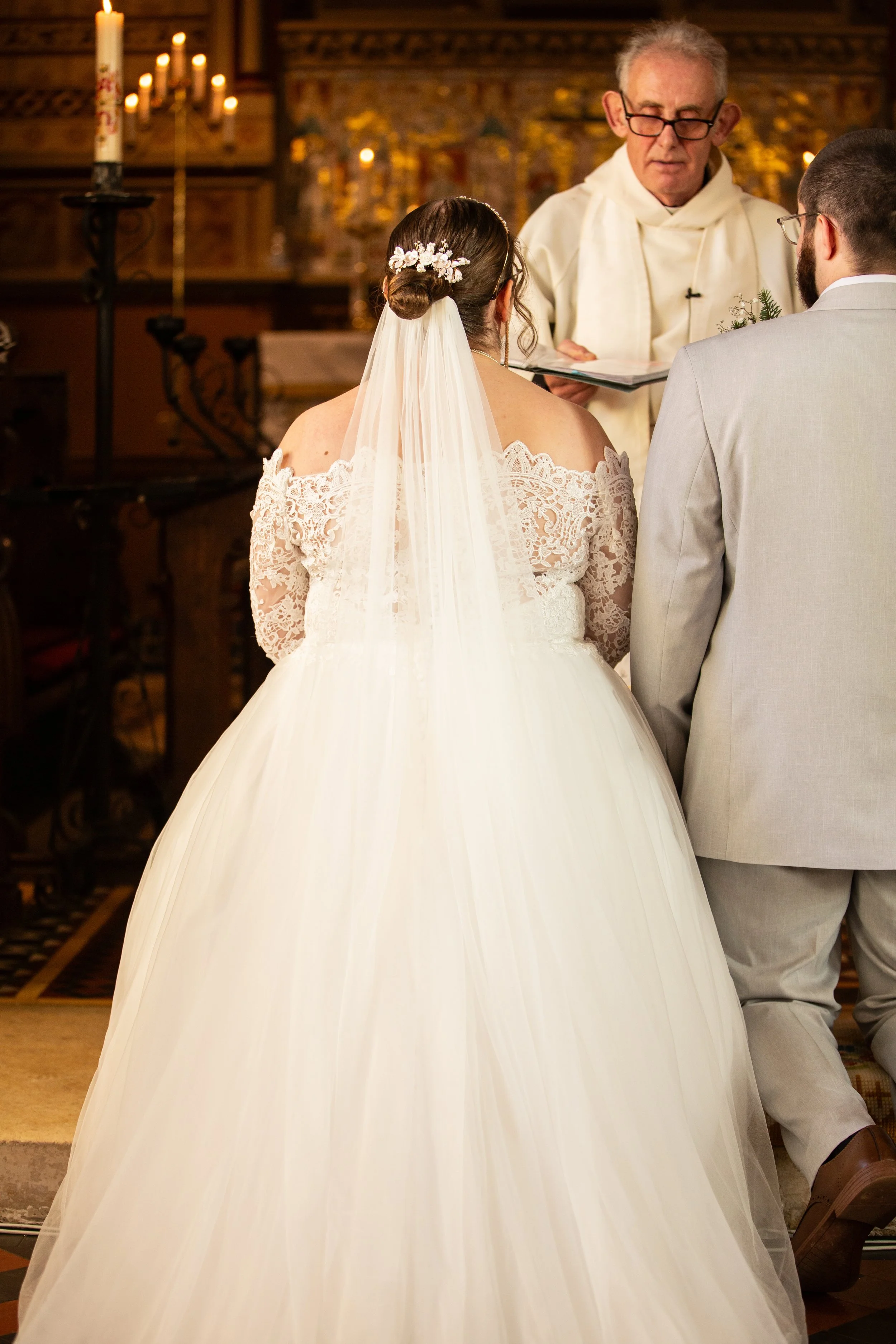 A bride and groom standing before a priest in a church during their wedding ceremony, with the focus on the bride's flowing white gown and lace off-shoulder sleeves, while the priest reads from a book.