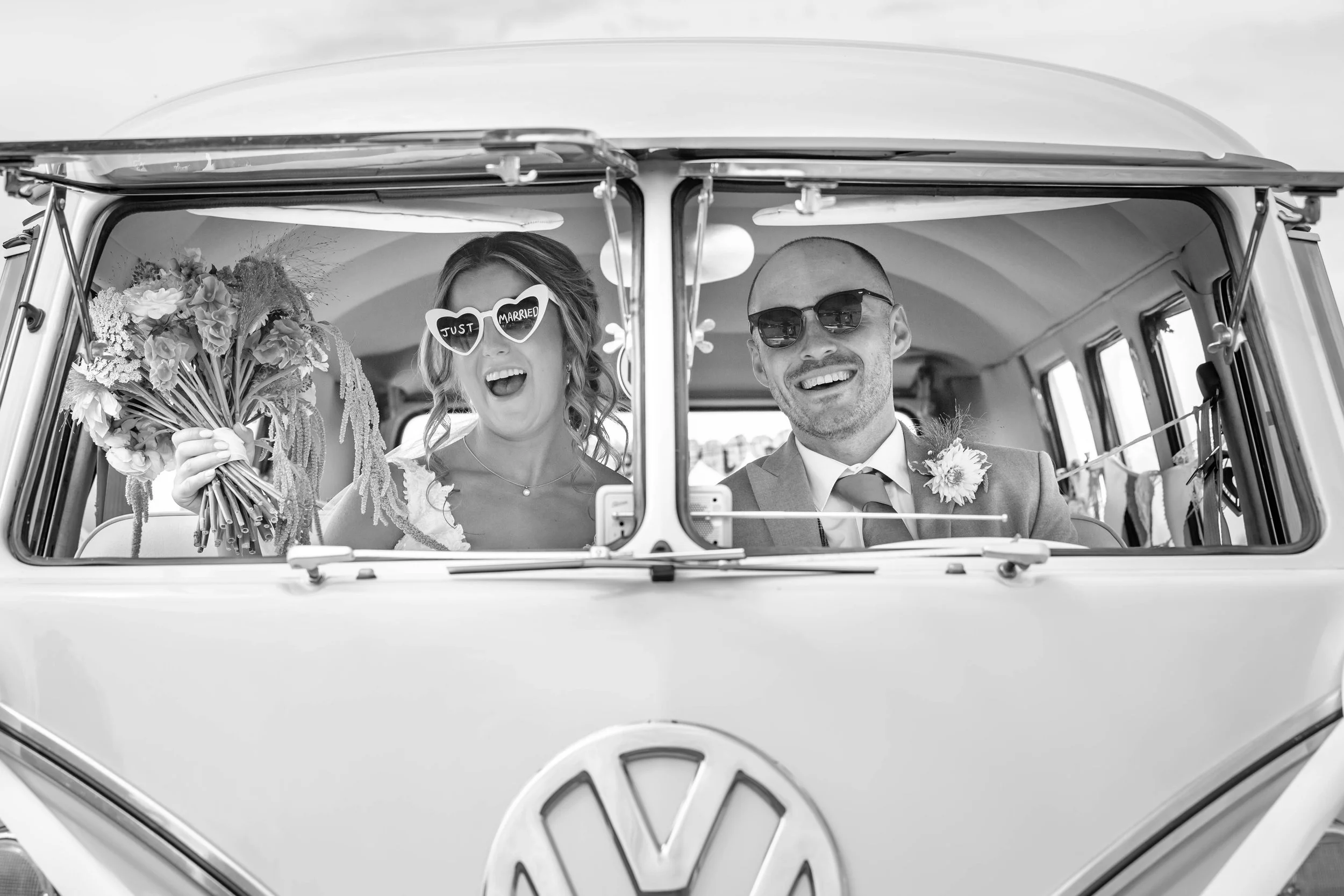 A happy couple in wedding attire inside a vintage Volkswagen bus, with the woman holding a bouquet and wearing heart-shaped sunglasses that say 'Just Married.'