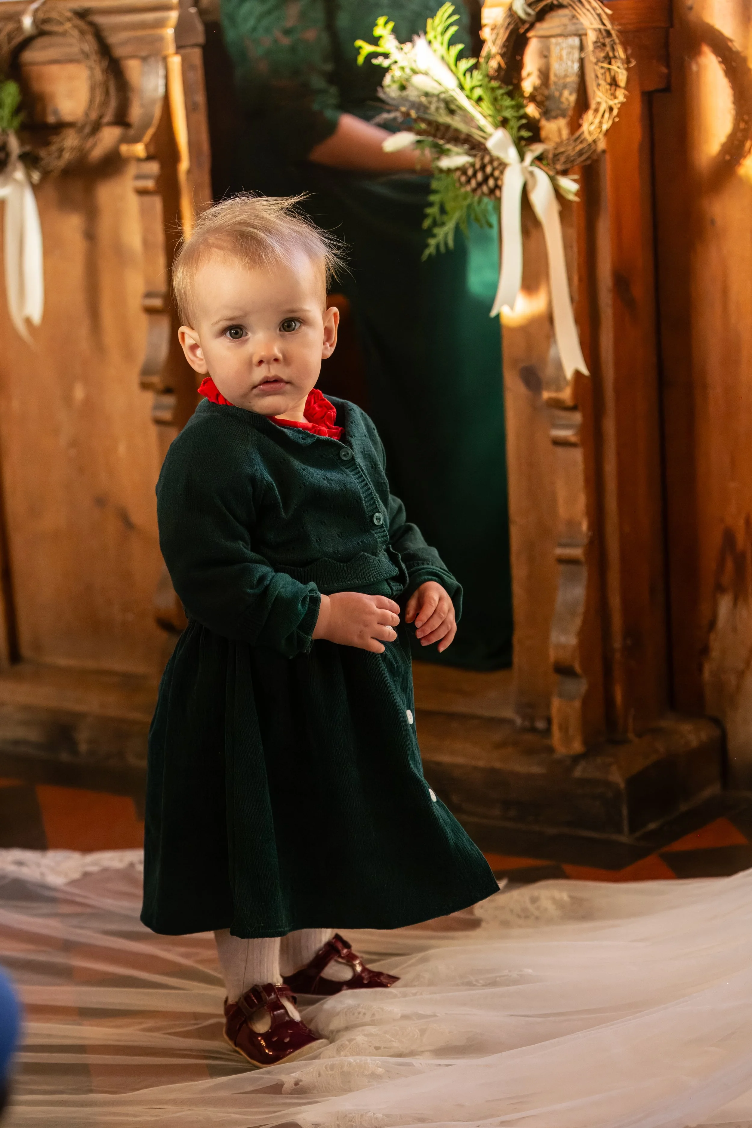 A young girl standing inside a wooden church, dressed in a dark green dress with a red ruffled collar, white tights, and shiny red shoes. She is looking directly at the camera with a curious expression. In the background, there are wooden pews and ho