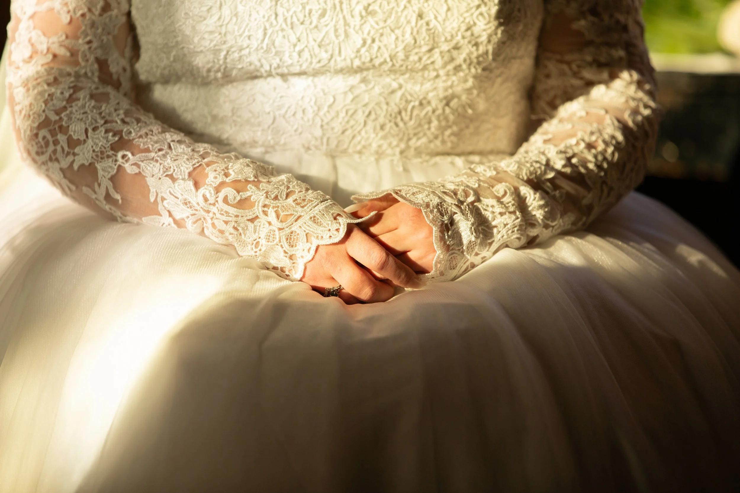 Close-up of a person in a cream lace dress sitting on a bed, with hands clasped on their lap, in natural light.