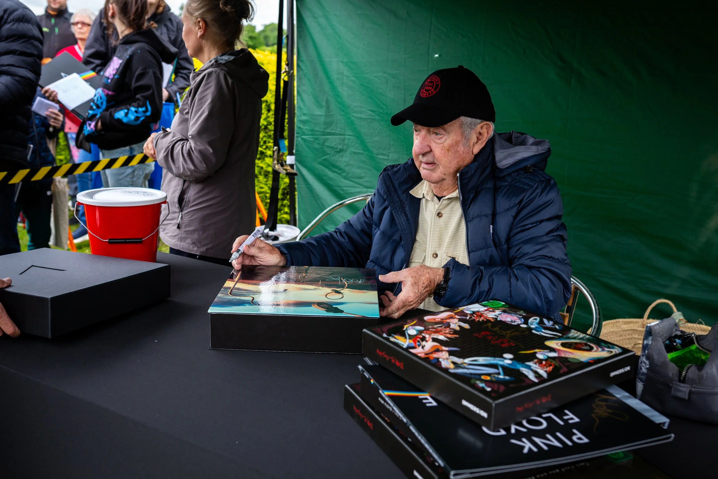 An elderly man wearing a black cap and a blue jacket signing a book at an event with people standing in line in the background.