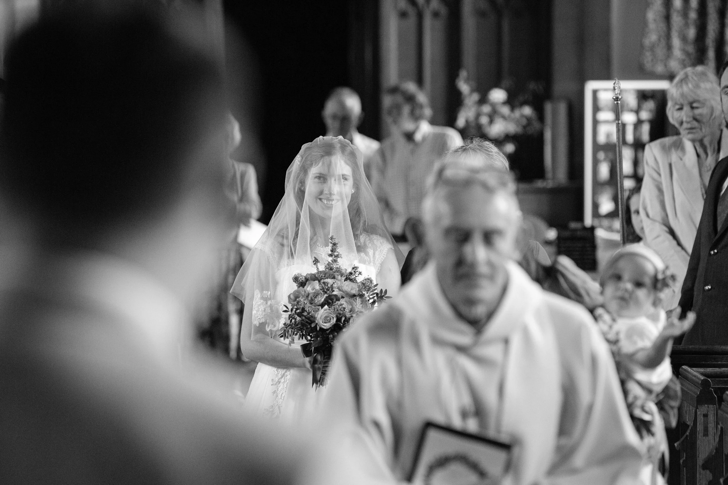 A bride smiling and holding a bouquet of flowers during a wedding ceremony, with guests seated around her in a church setting.