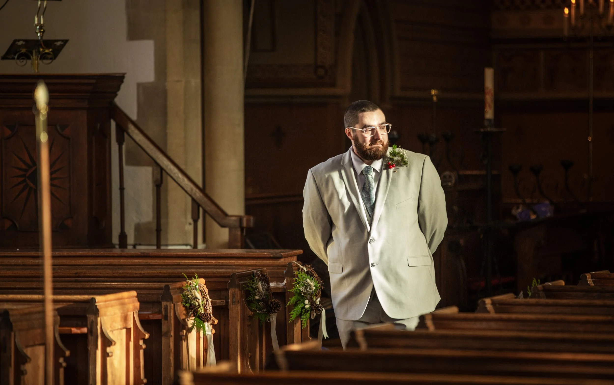 A man with glasses and a beard in a suit with a holiday decoration on his lapel stands inside a church with wooden pews and candleholders.