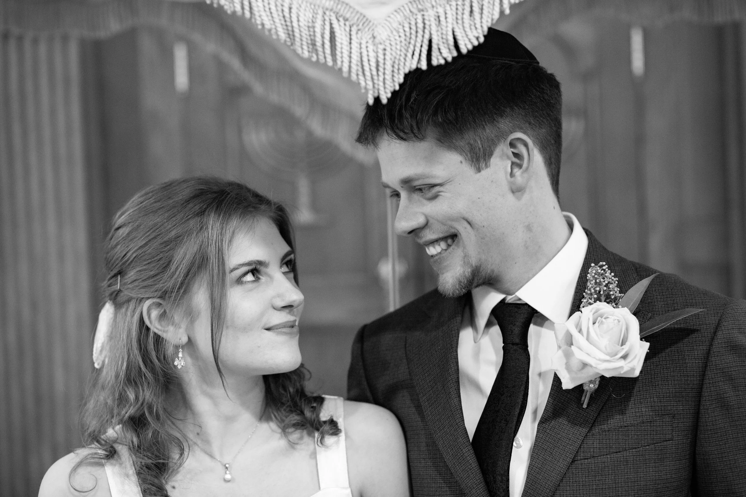A black and white photo of a bride and groom during their wedding. The bride is looking at the groom, who is smiling and looking down at her. The groom is wearing a suit with a boutonnière, and the bride is wearing earrings and a necklace.