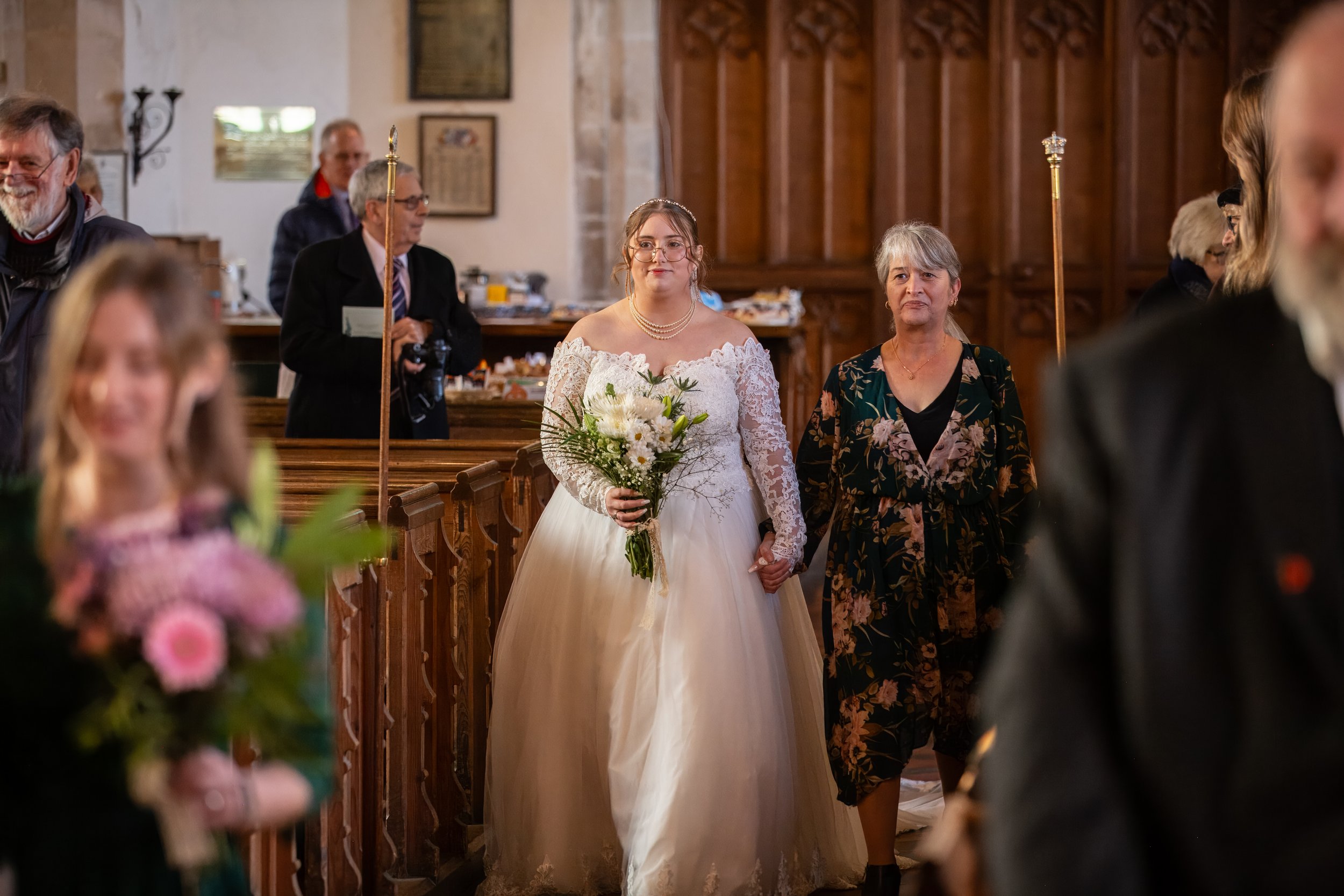 Bride in a white lace wedding dress and pearl necklace holding a bouquet of white flowers, standing in a church aisle next to a woman in a floral dress during a wedding ceremony.