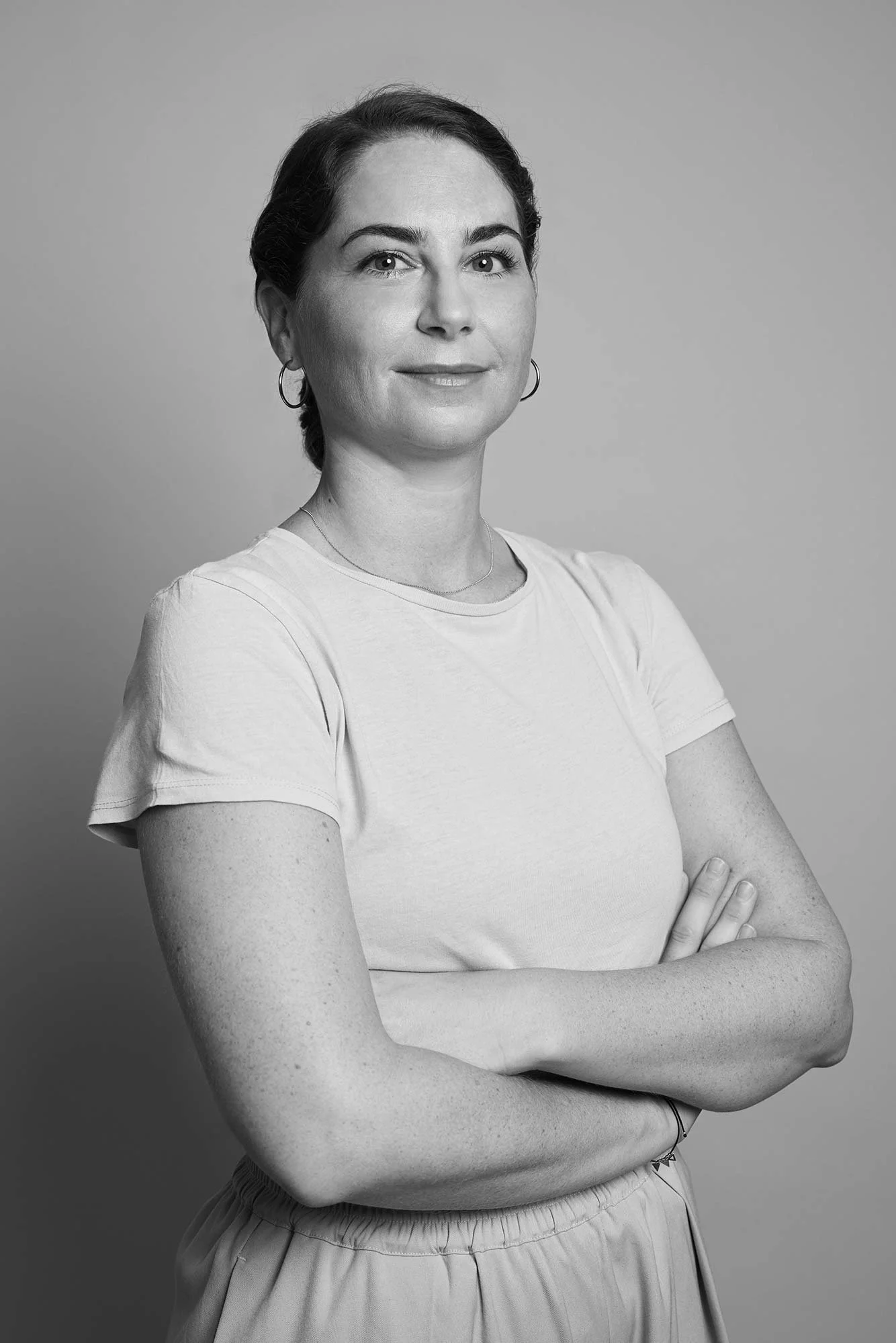 Black and white portrait of a woman with crossed arms, smiling slightly, wearing a light T-shirt, earrings, and a necklace, against a plain background.