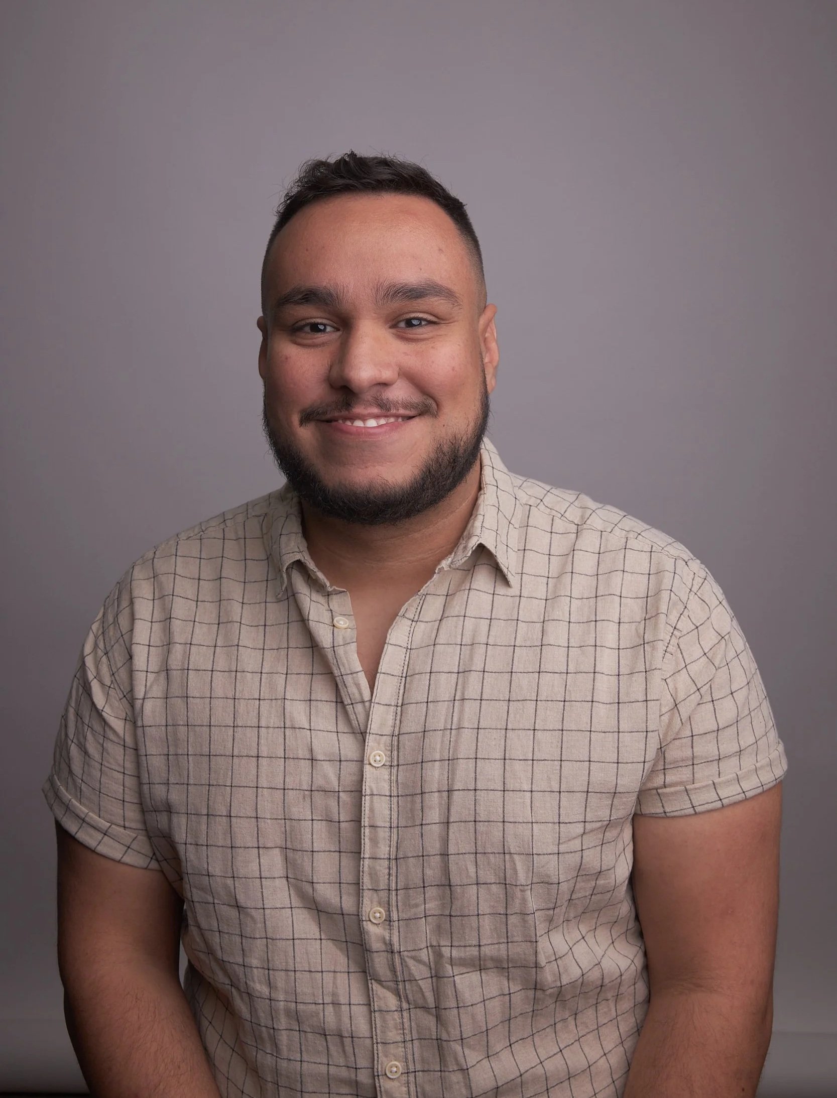 Portrait of a young man with dark hair, beard, and light skin, smiling, wearing a checked short-sleeve button-up shirt, against a plain gray background.