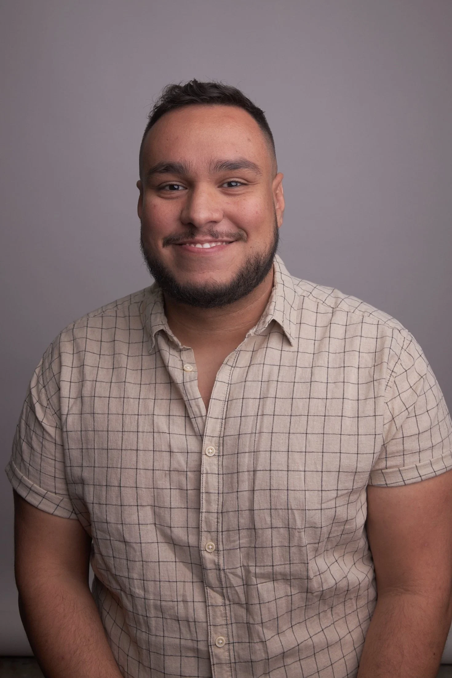 A young man with a beard and short dark hair, wearing a beige checkered shirt, smiling at the camera against a plain gray background.