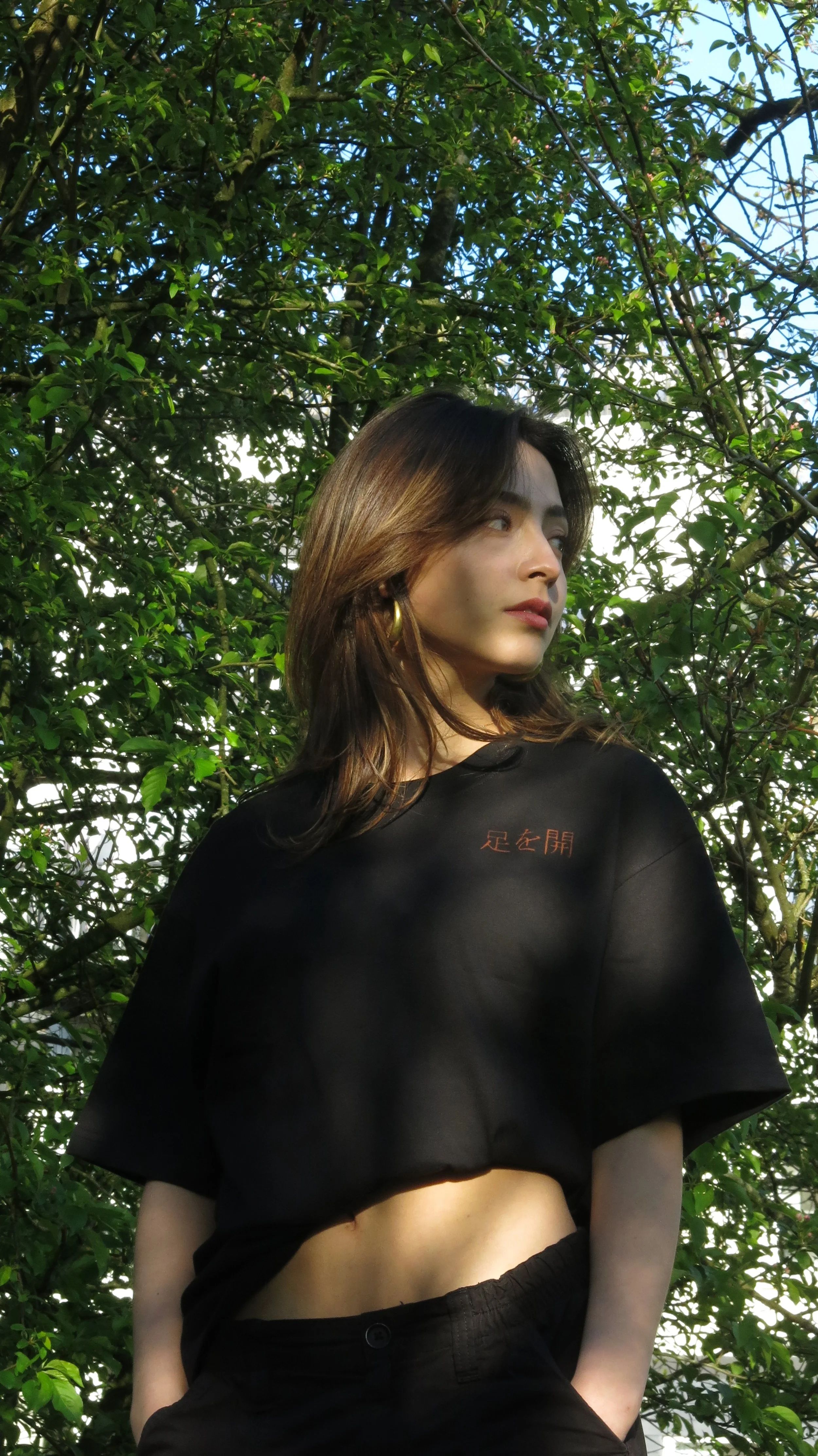A young woman with long brown hair and gold hoop earrings standing outdoors among green leafy trees, wearing a black crop top with red Japanese characters on it, looking off to the side.