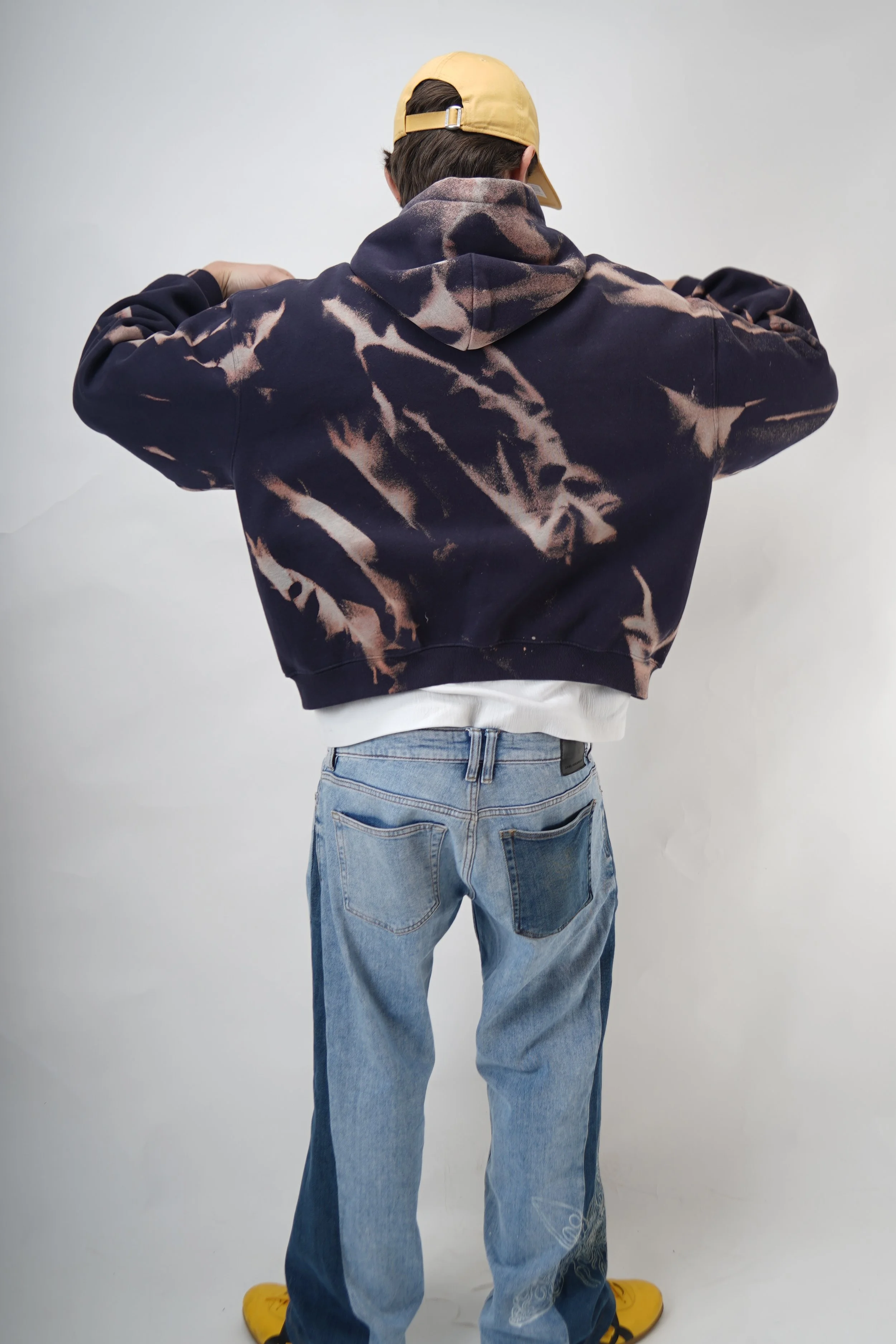 A person standing with their back facing the camera, wearing a beige cap, a dark tie-dye hoodie, light blue jeans, and yellow shoes.