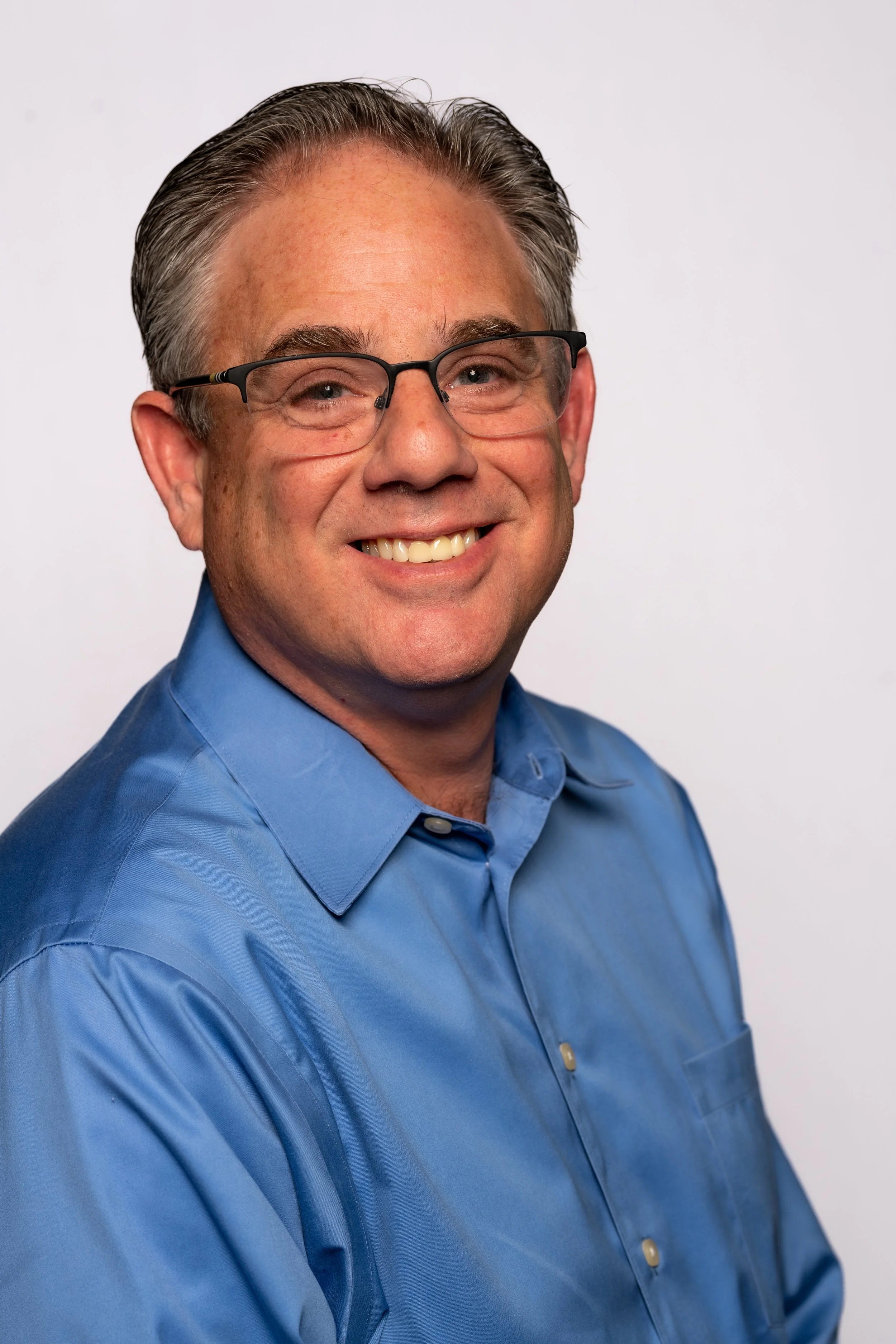 A middle-aged man with short gray hair, wearing glasses, a blue dress shirt, and smiling, against a plain white background.