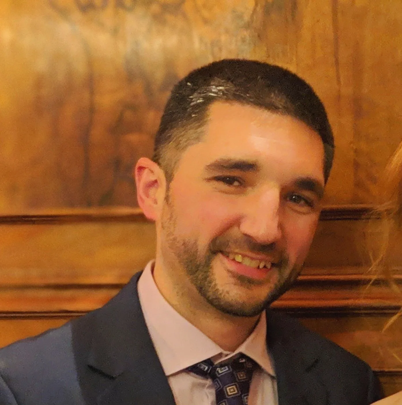 A man with dark hair and a beard, wearing a suit and tie, smiling in front of a wooden background.