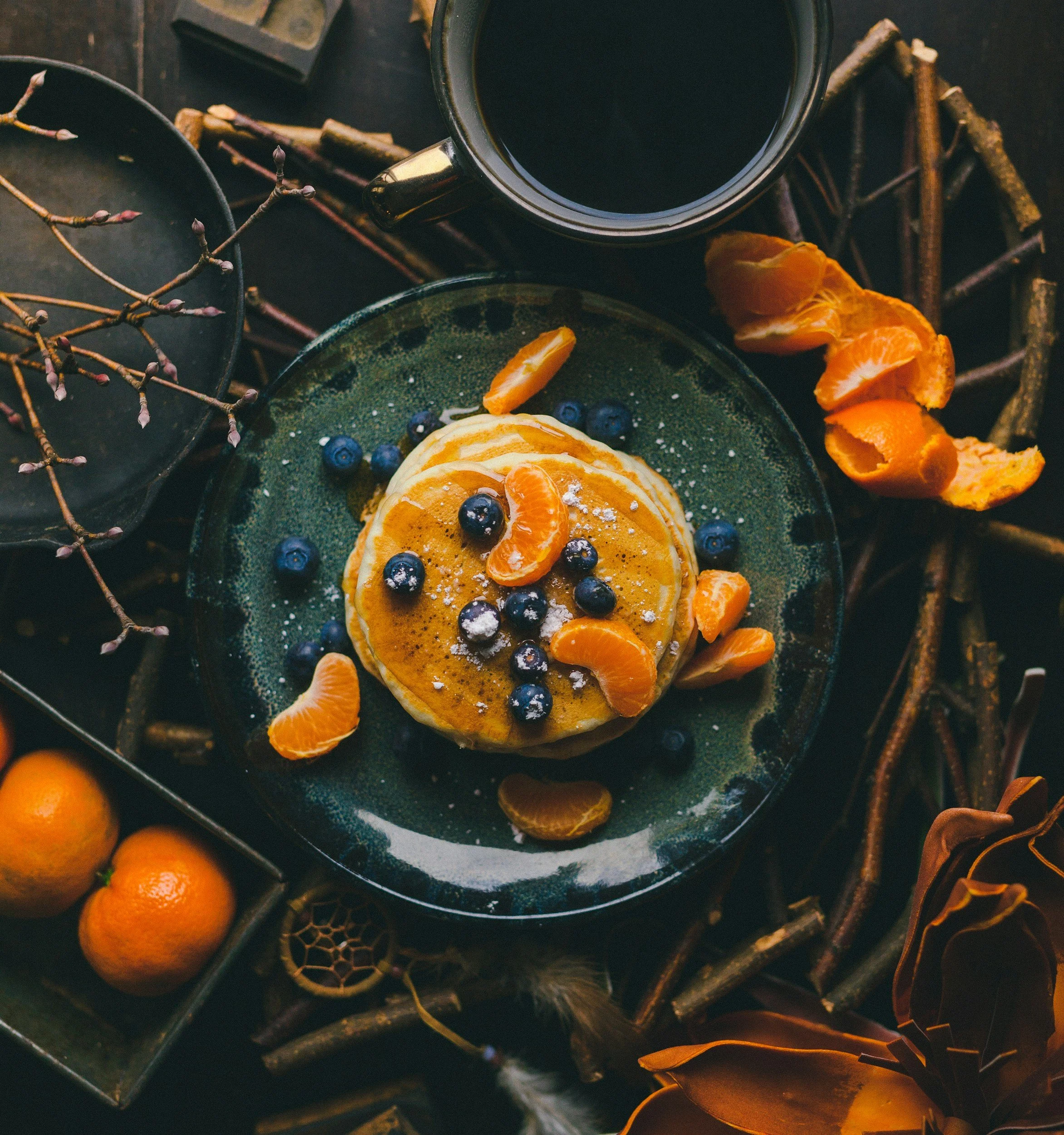 A top-down view of a breakfast scene with a stack of pancakes topped with blueberries and orange slices, accompanied by a cup of black coffee, surrounded by orange and purple berries, twigs, and decorative elements on a dark surface.