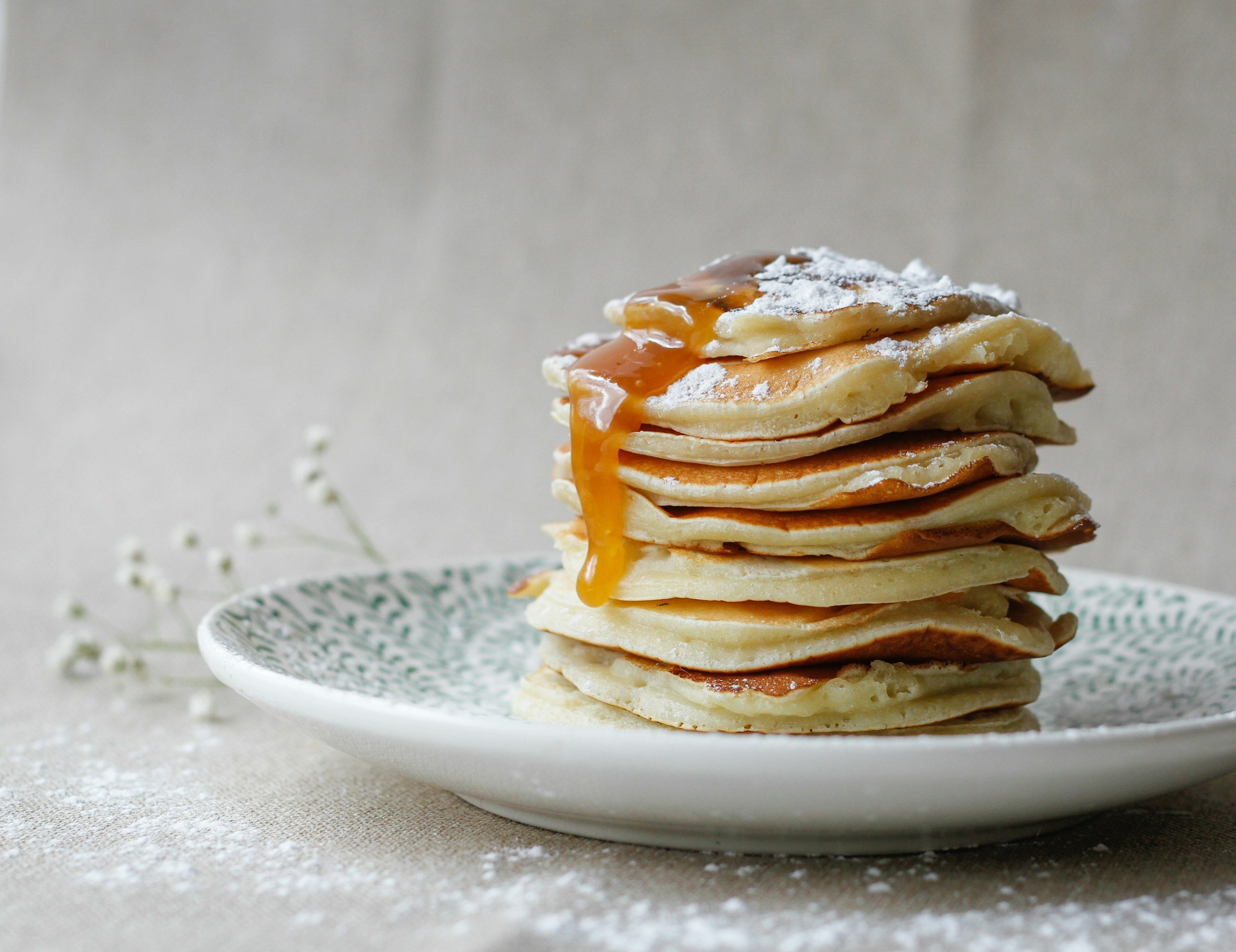 Stack of five pancakes with caramel sauce and powdered sugar on top, served on a white patterned plate with a grey background.