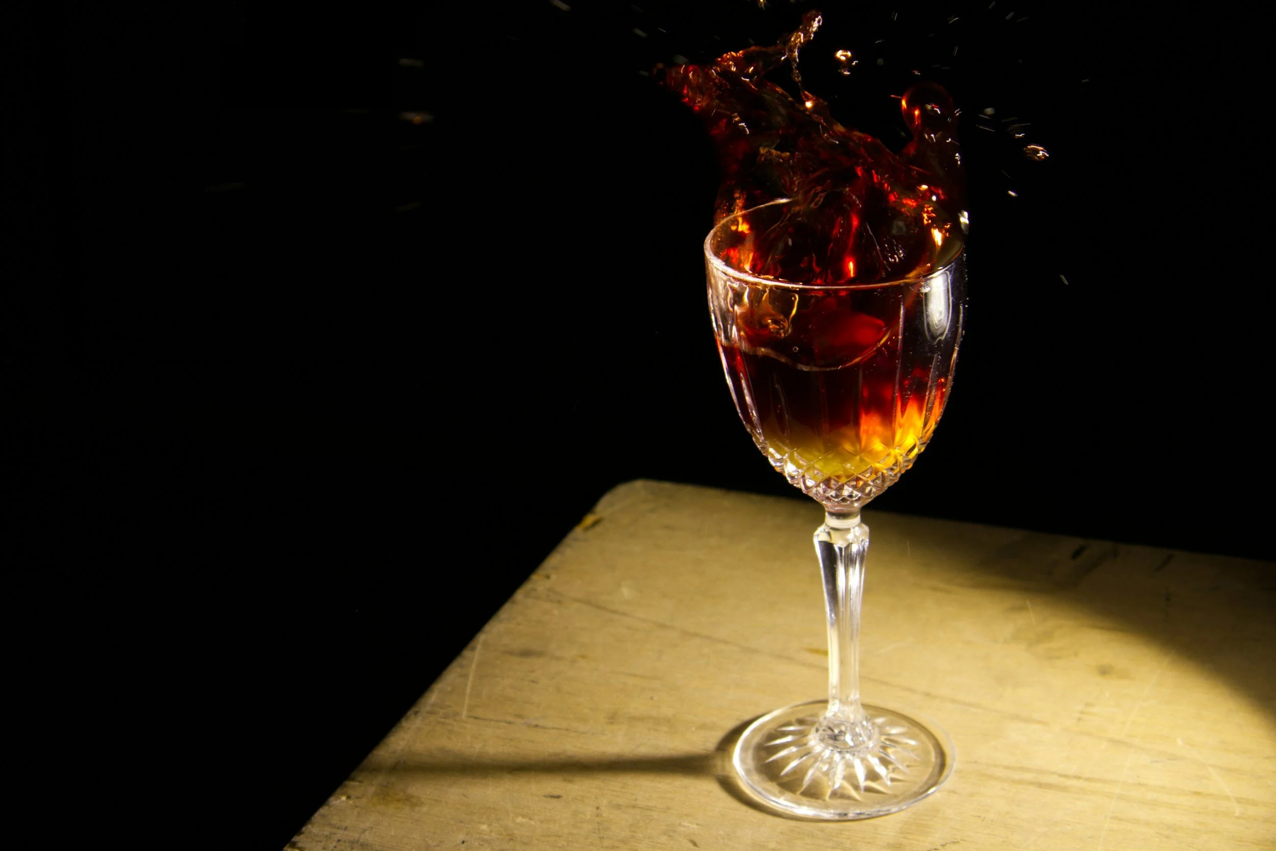 A glass of red liquid, possibly wine, with liquid splashing out, placed on a wooden surface against a black background.
