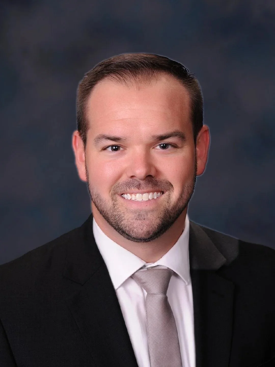 Professional headshot of a smiling man wearing a black suit, white shirt, and beige tie, against a dark, blurred background.