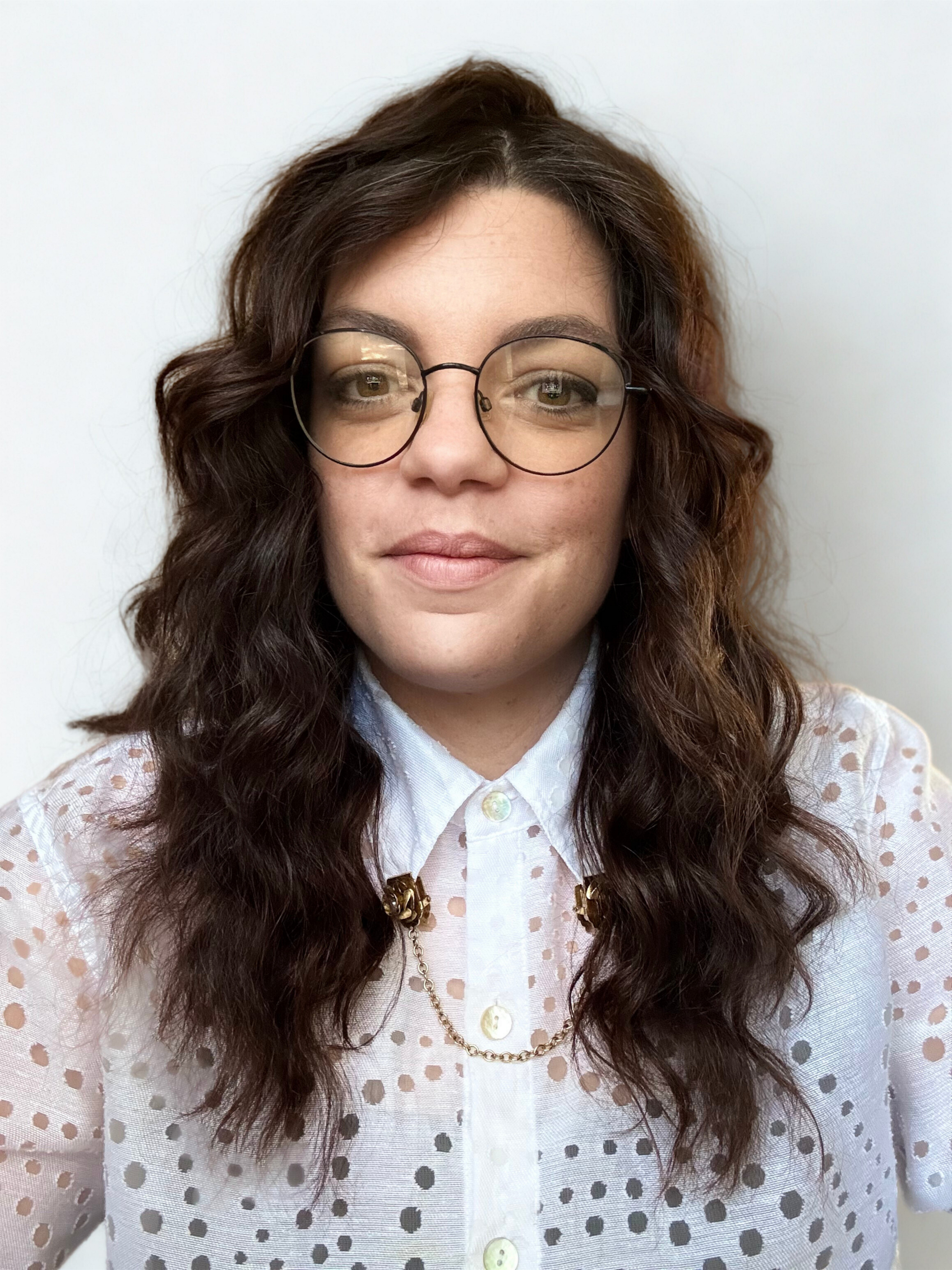 A woman with long, curly brown hair wearing glasses, a white polka-dot blouse, and gold jewelry, standing against a plain white background.