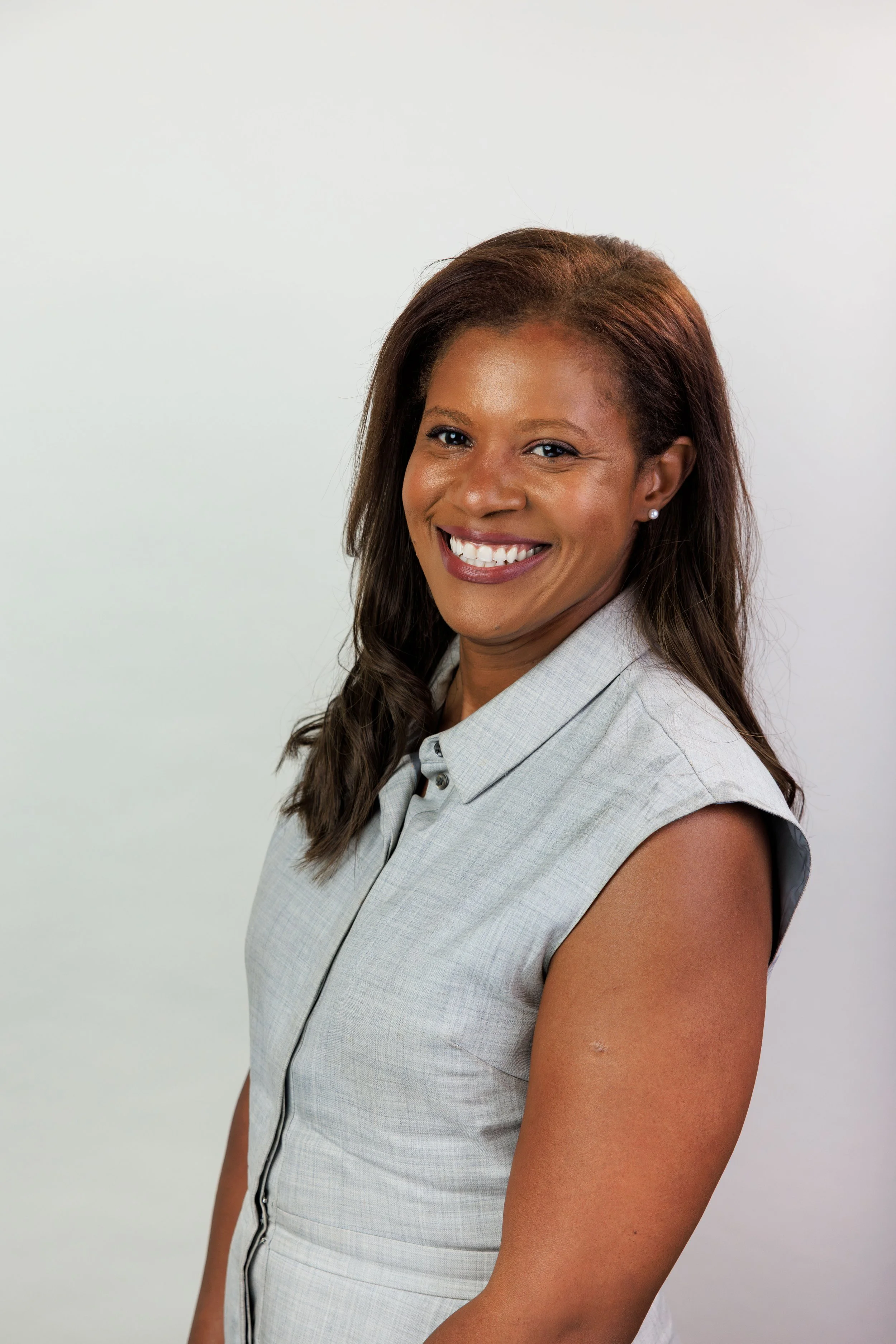 A smiling woman with brown skin, long curled hair, wearing a sleeveless gray plaid blazer, standing against a plain white background.