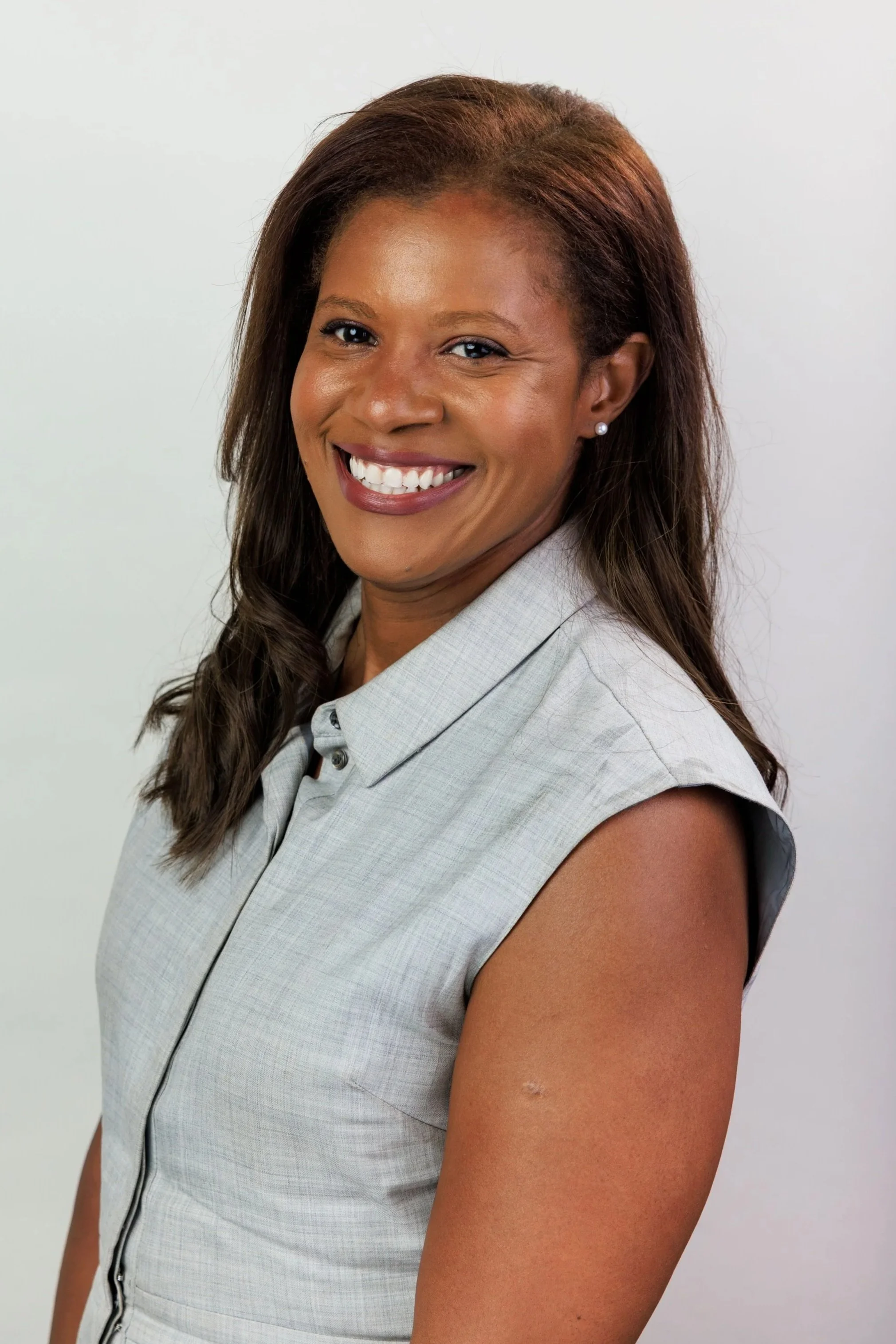 A smiling African American woman with long brown hair, wearing a sleeveless light gray button-up shirt and pearl earrings, standing against a plain white wall.