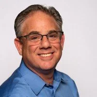 A smiling man with glasses, short gray hair, wearing a blue button-up shirt, against a plain white background.