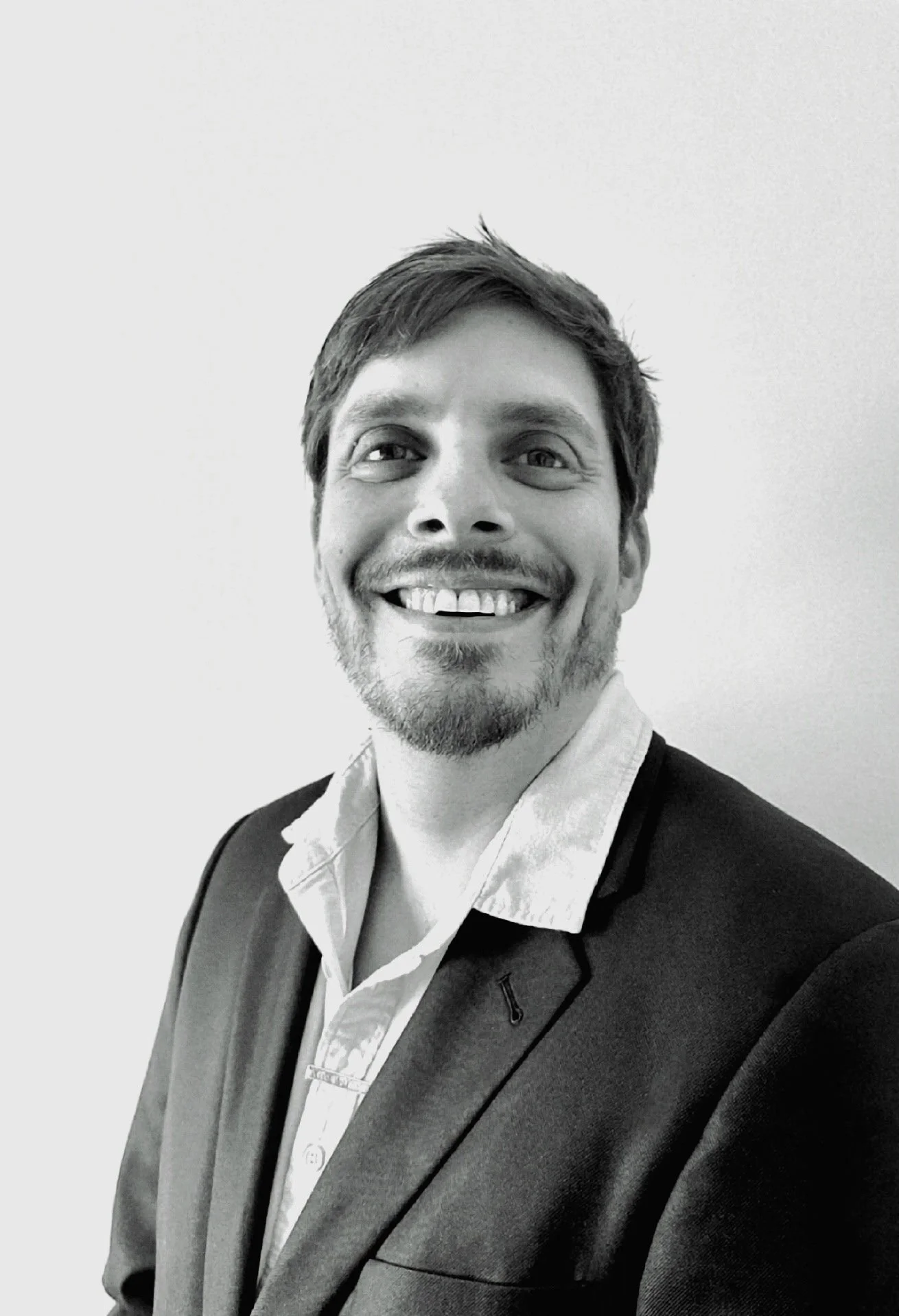 Black and white photo of a man wearing a suit, white shirt, and backpack. He is smiling and looking slightly to his left.