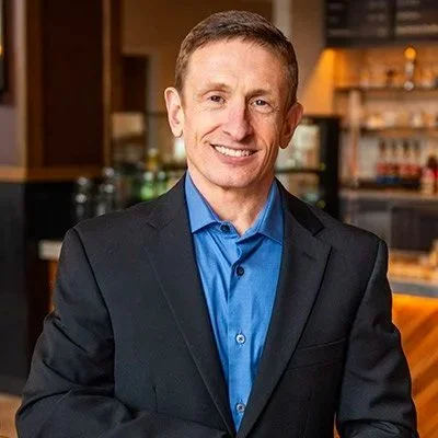 A man in a dark suit and blue shirt smiling in a kitchen or bar area.