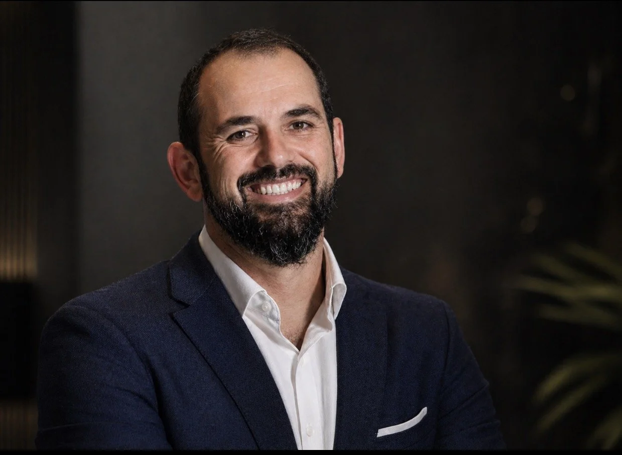 A smiling man with dark hair, a beard, and a mustache, wearing a navy blazer over a white dress shirt, standing against a dark background.