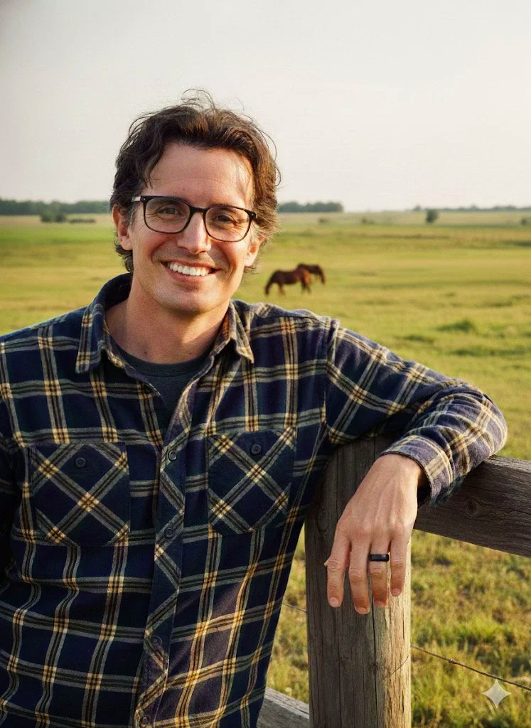A man with glasses and dark wavy hair smiling, leaning on a wooden fence in a grassy field with a horse grazing in the background during daytime.