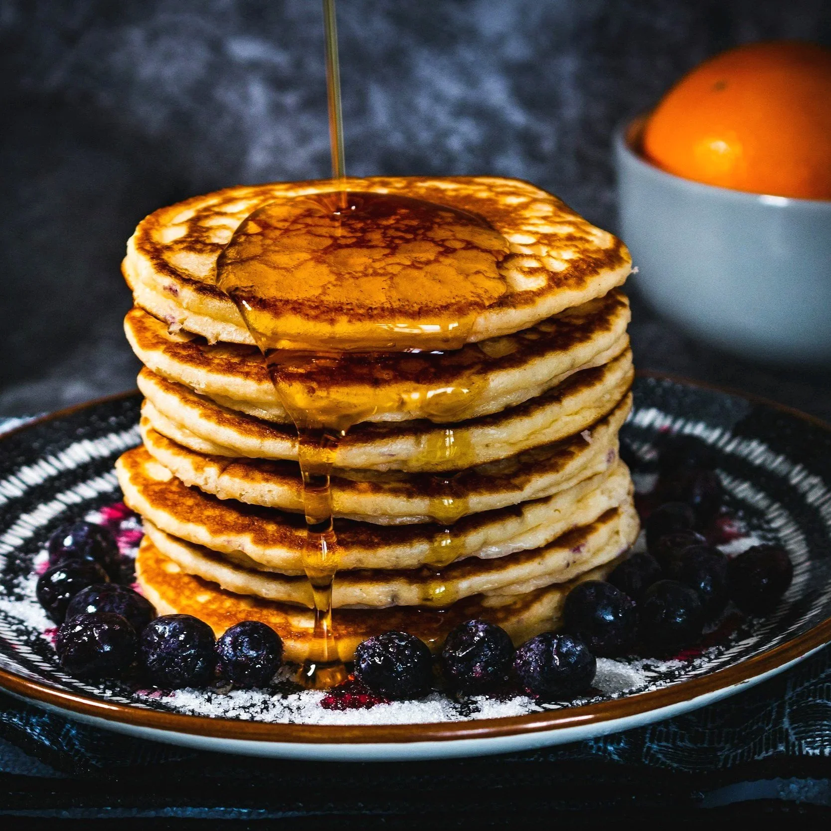 Stack of four golden-brown pancakes topped with syrup, surrounded by blueberries on a black-patterned plate.