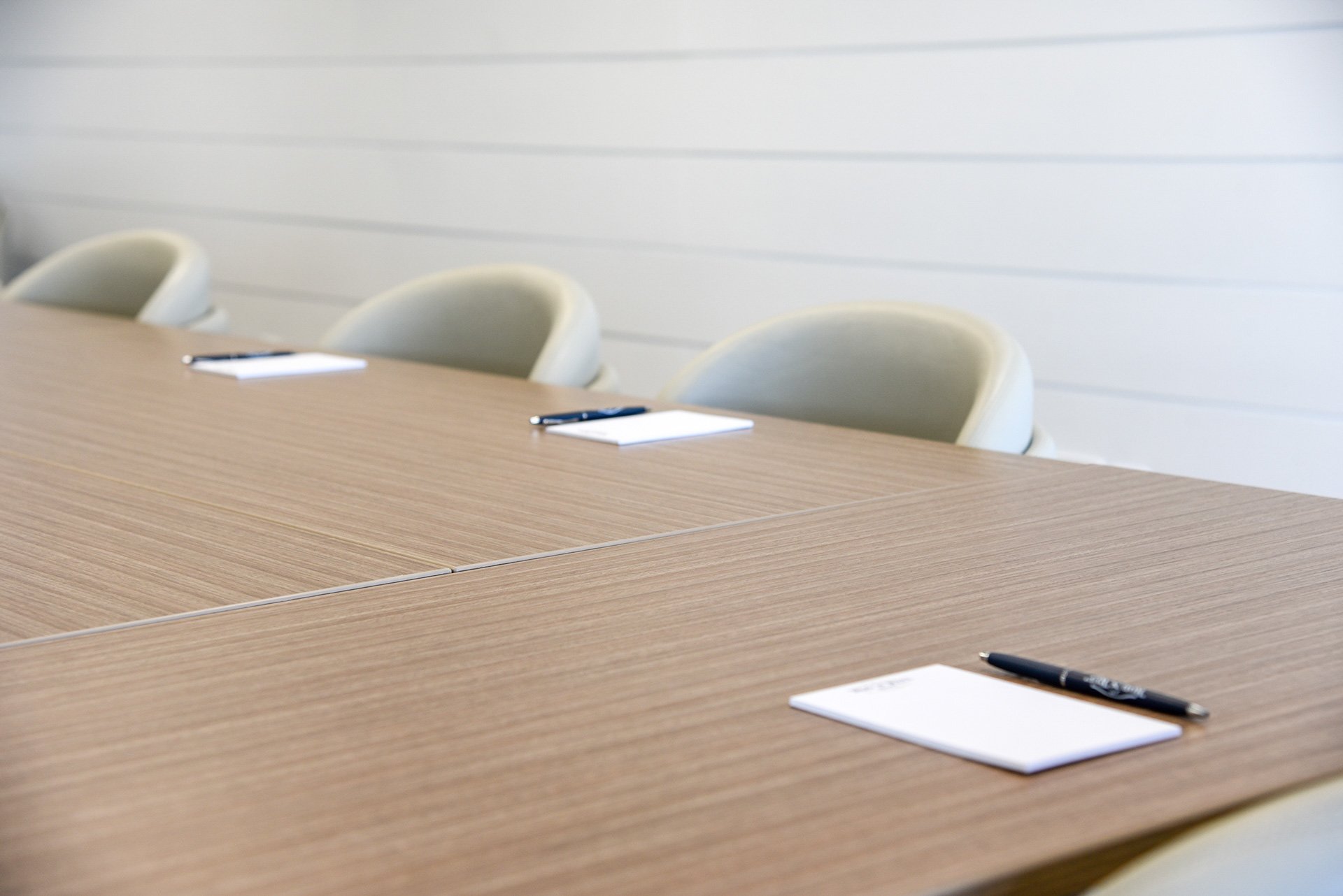 Conference room table with notepads and pens, beige chairs against a white wall.