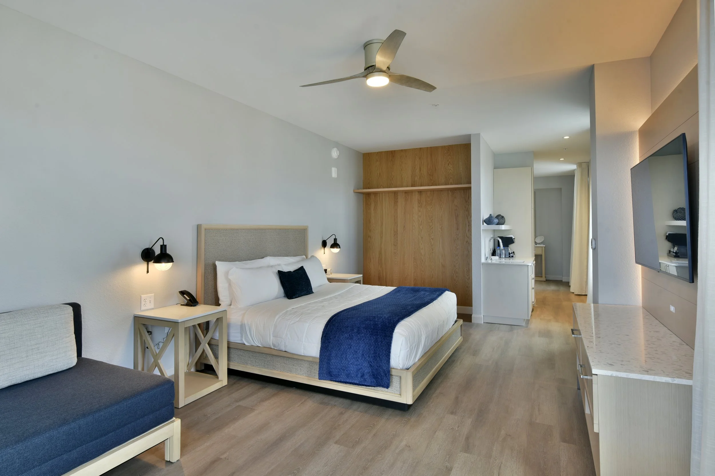Modern hotel room with a bed, nightstand, lamps, sofa, wooden panel accent wall, flat-screen TV, kitchenette, and wooden flooring.