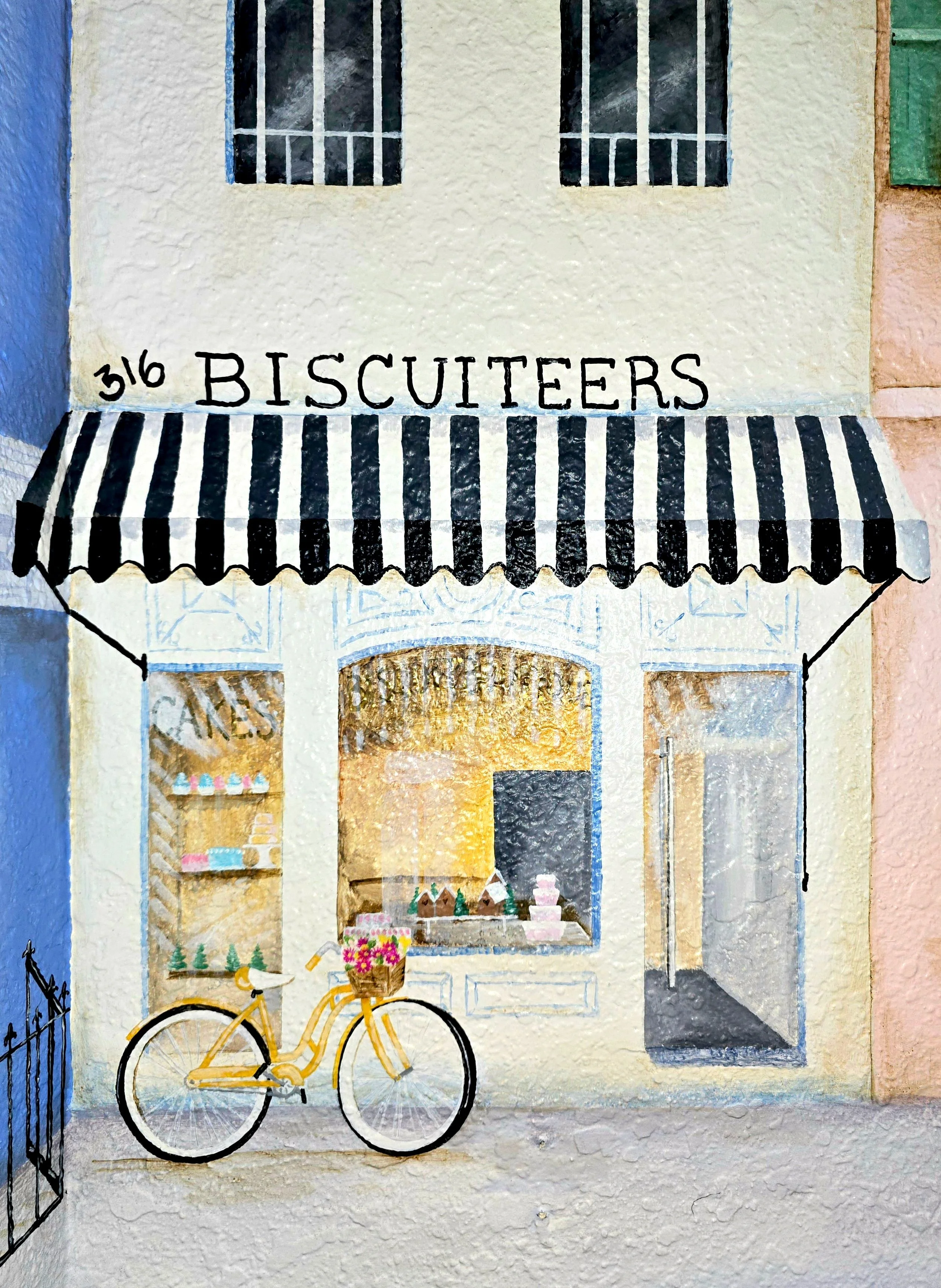 A pastel-colored storefront with a black and white striped awning, two upper windows with bars, a yellow bicycle with a basket of flowers parked outside, and a display window with baked goods inside. The store's name is 'Biscuitters' and the address is 316.