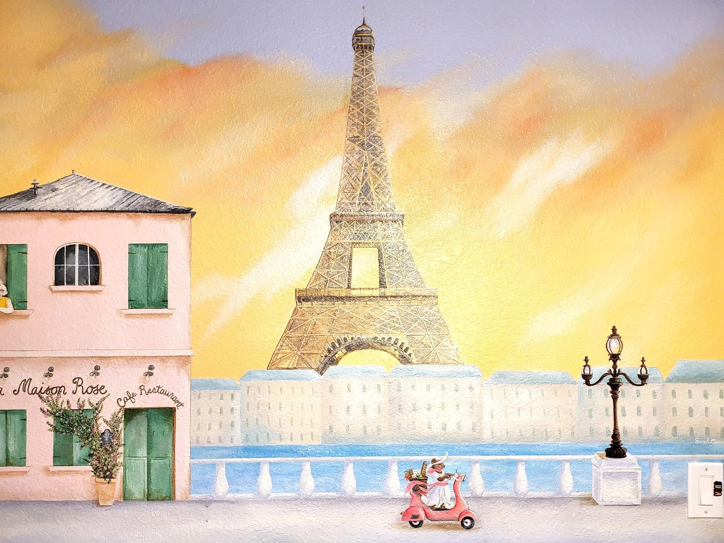 A wall mural of Paris featuring the Eiffel Tower, a pink building with green shutters, a vintage street lamp, and a woman riding a pink scooter in front of the Seine River during sunset.