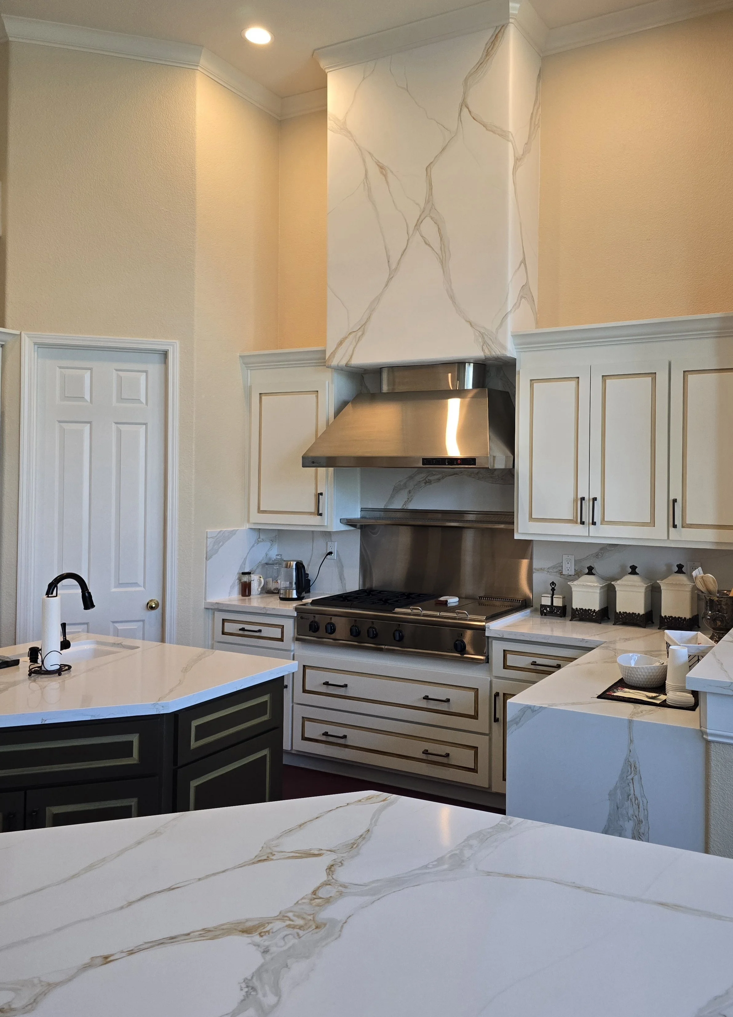 Modern kitchen with white marble countertops, stainless steel stove and hood, and white cabinets with gold trim.
