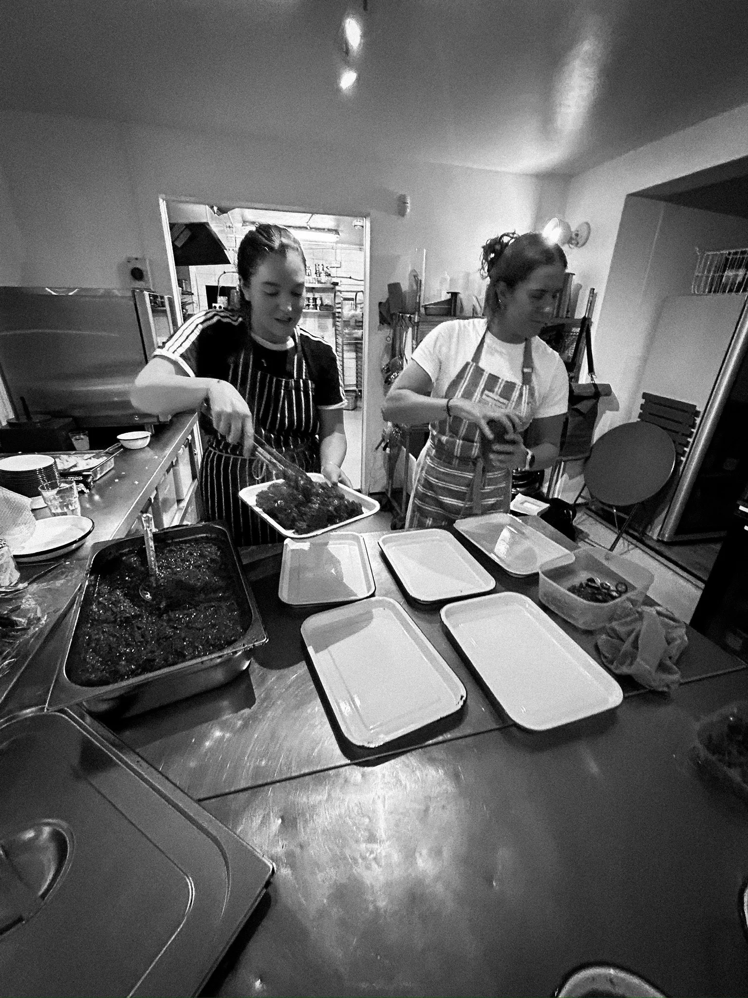 Two women preparing food in a kitchen, with trays and ingredients on the counter.