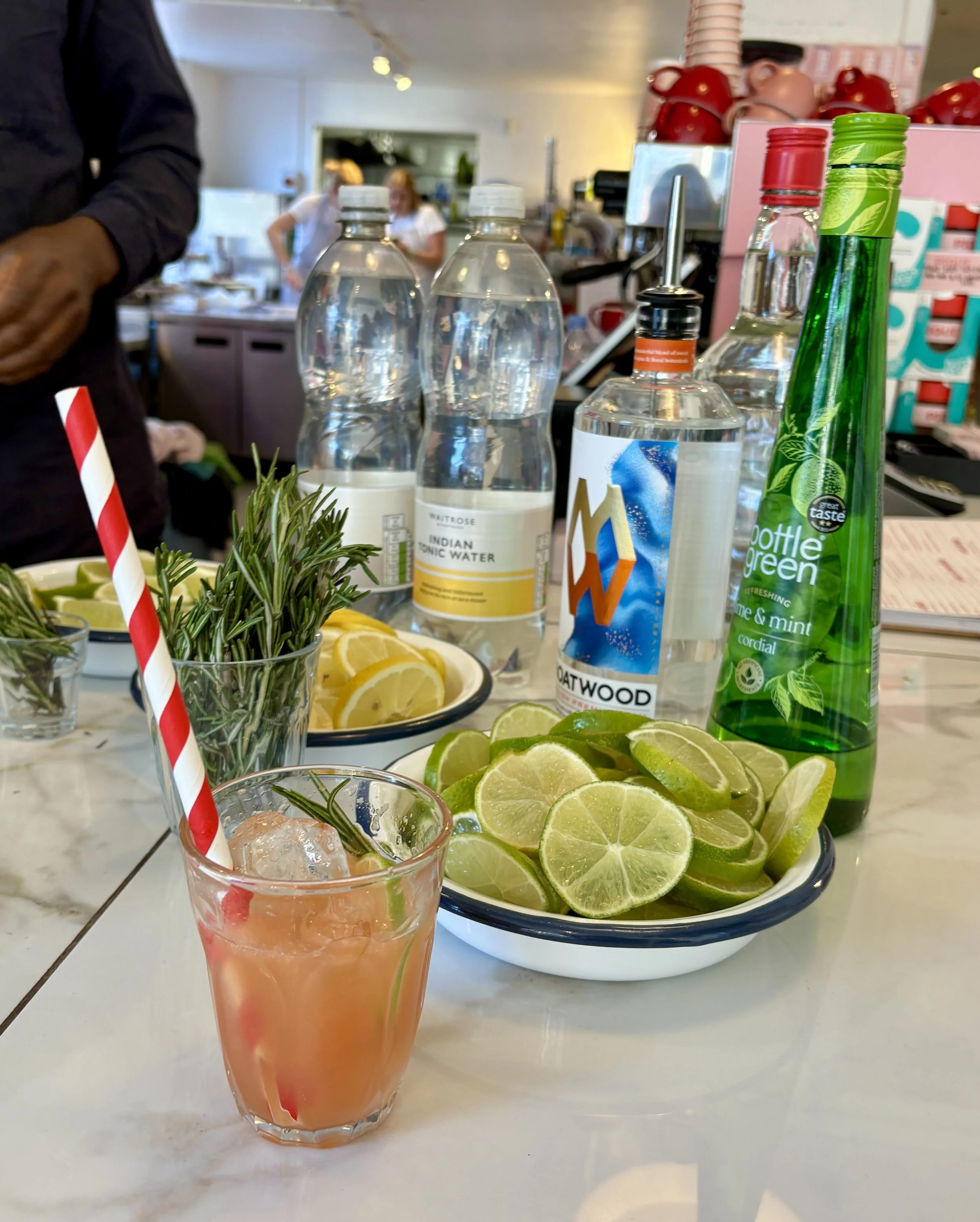 Bar counter with lime slices, lemon slices, rosemary sprigs, bottles of tonic water, flavored sparkling water, and a pink cocktail with ice and a striped straw.