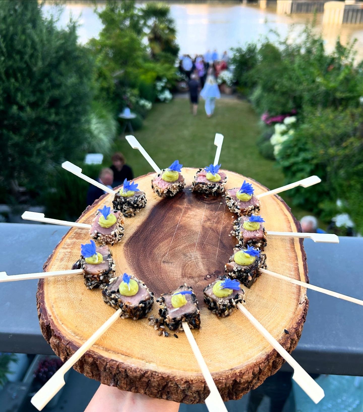 A wooden serving platter with small appetizers topped with yellow and blue garnishes, arranged in a circle, with a garden and guests in the background.