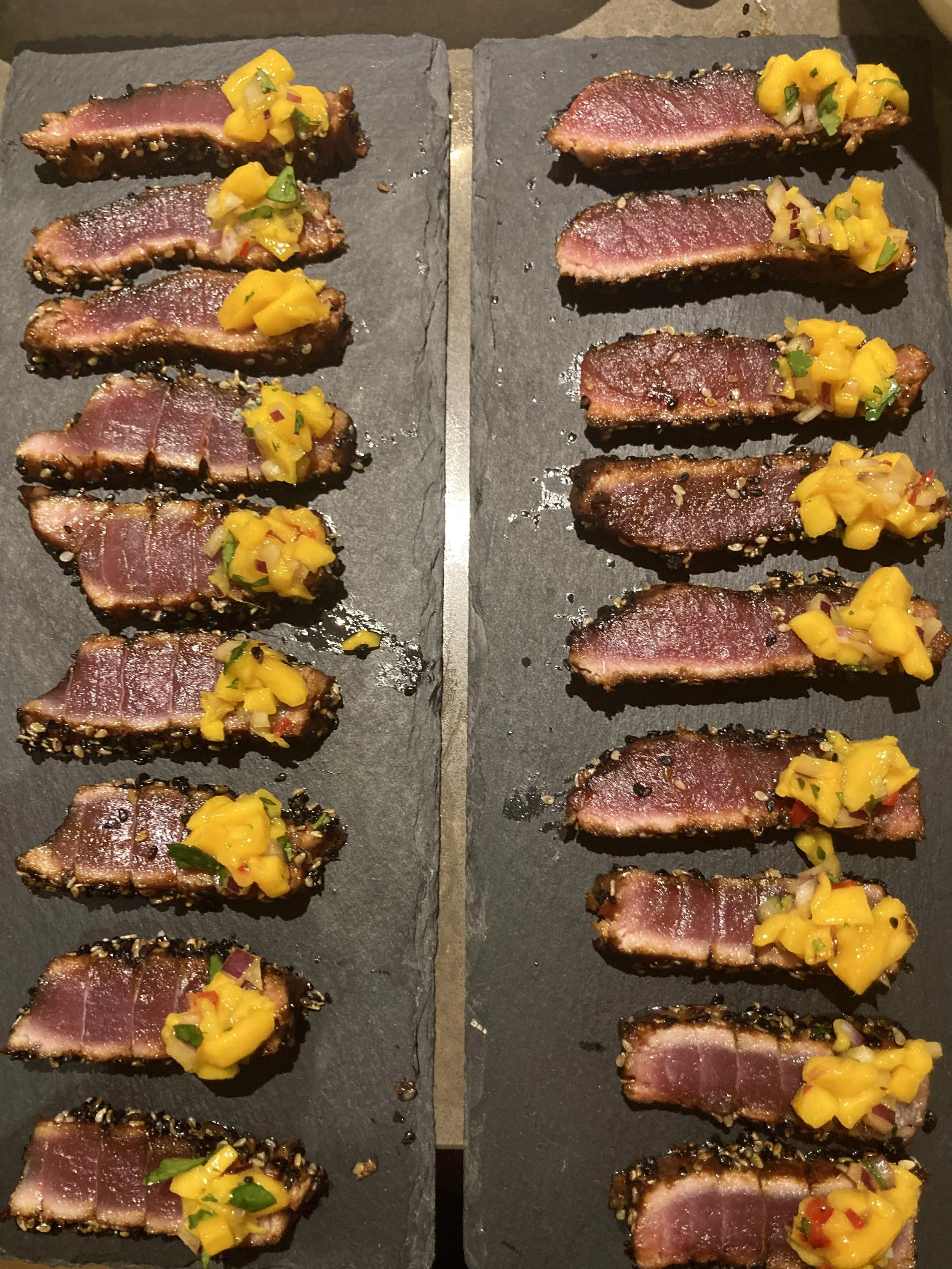 Slices of seared tuna with sesame seeds on a black slate serving board, topped with a mango salsa.