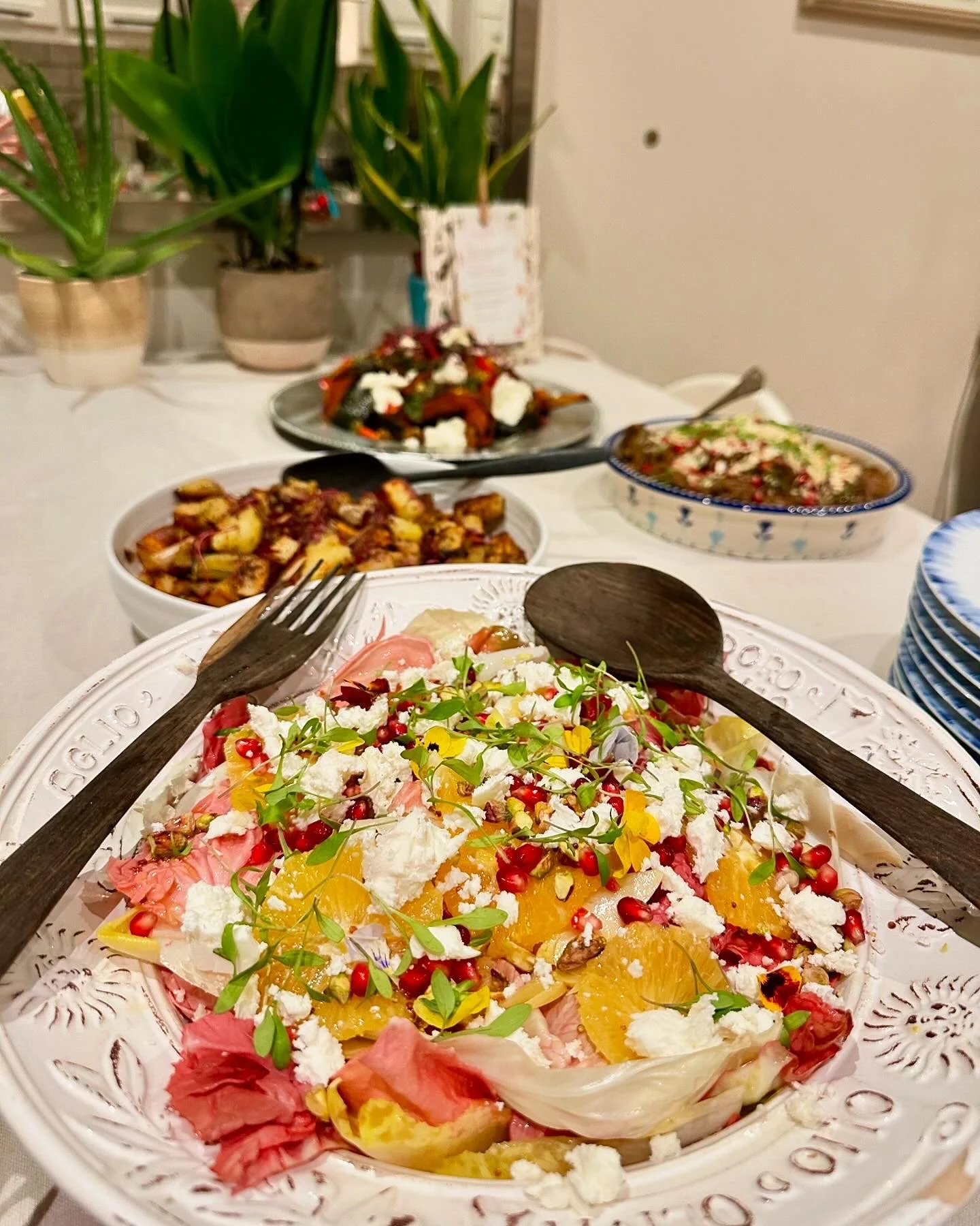 Colorful salad with pomegranate seeds, orange slices, feta cheese, and microgreens on a decorative white plate with wooden utensils, with various other dishes and potted plants visible in the background.