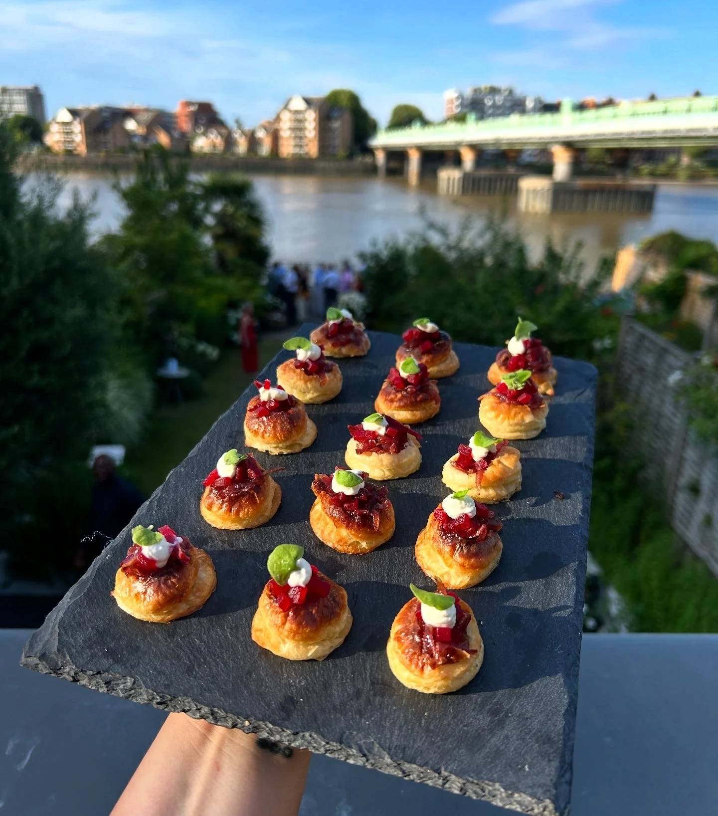 Close-up of a slate serving platter with small appetizers topped with red sauce, green herbs, and white dollops, held outdoors by a person's hand with a river and urban buildings in the background.