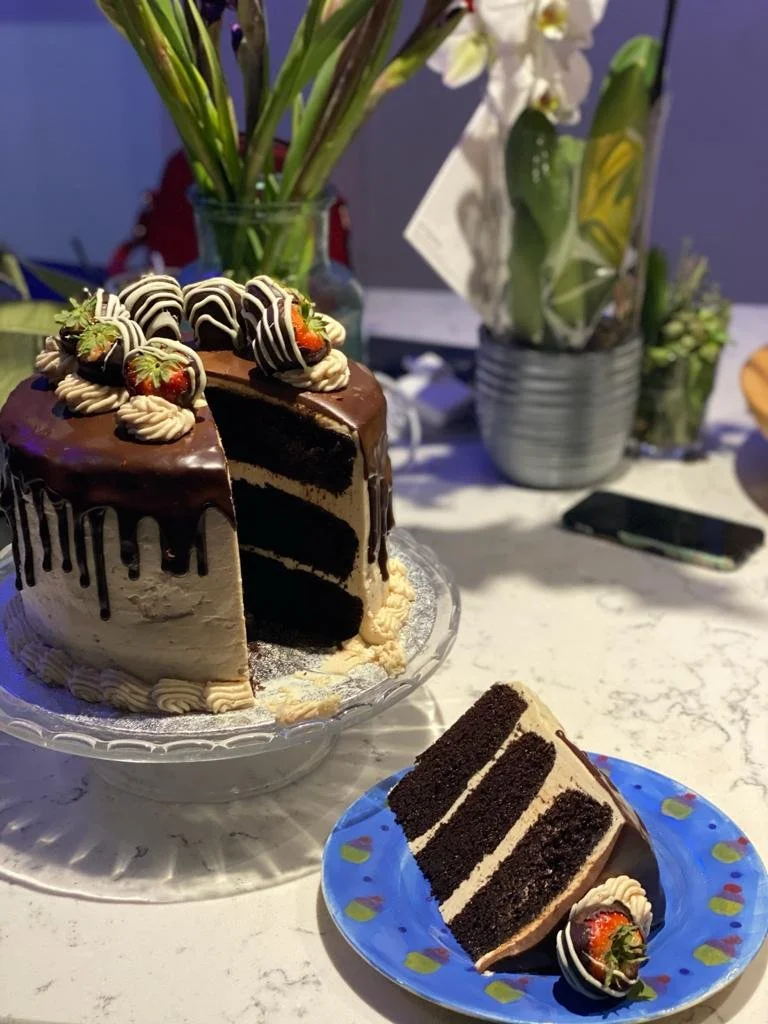Chocolate layered cake with strawberries and whipped cream on top, sliced to show layers, served on a blue plate with a decorative border, next to a slice of the same cake on a glass cake stand, with floral arrangements in the background.
