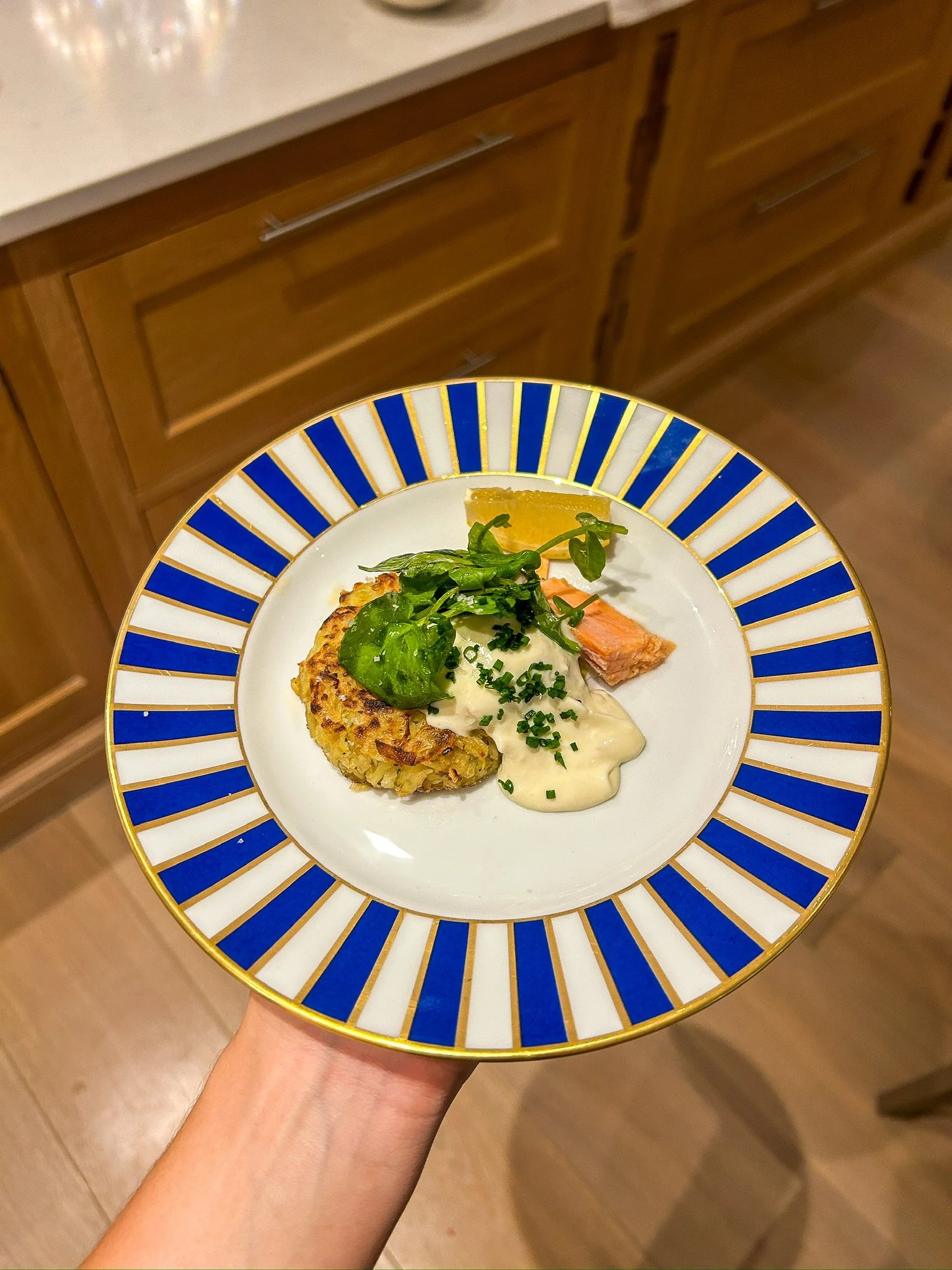 A decorative plate with a blue, white, and gold border holds a serving of salmon, a potato pancake topped with chives, a dollop of white sauce, a slice of lemon, and a small green salad with arugula and herbs.