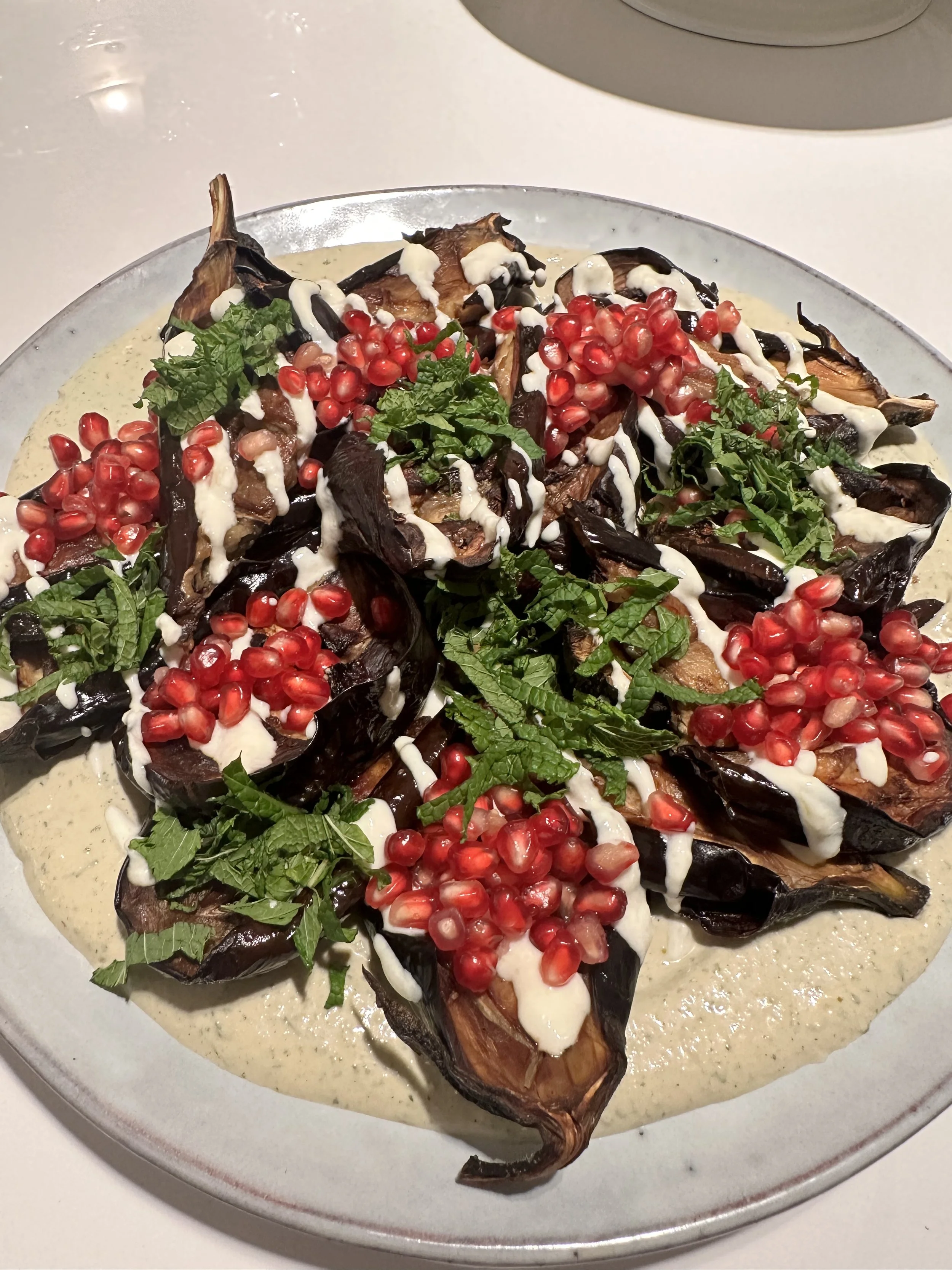 A plate of roasted eggplant topped with pomegranate seeds, fresh herbs, and drizzled with sauce over a creamy greenish sauce.