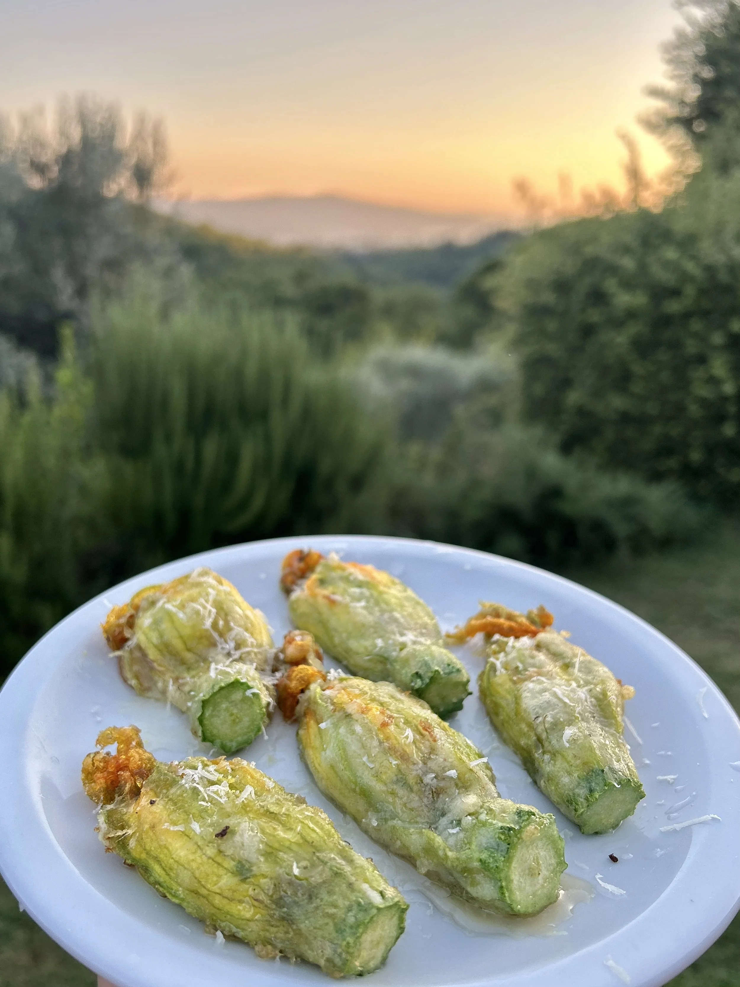 Fried green chilies on a white plate with a scenic sunset and greenery in the background.