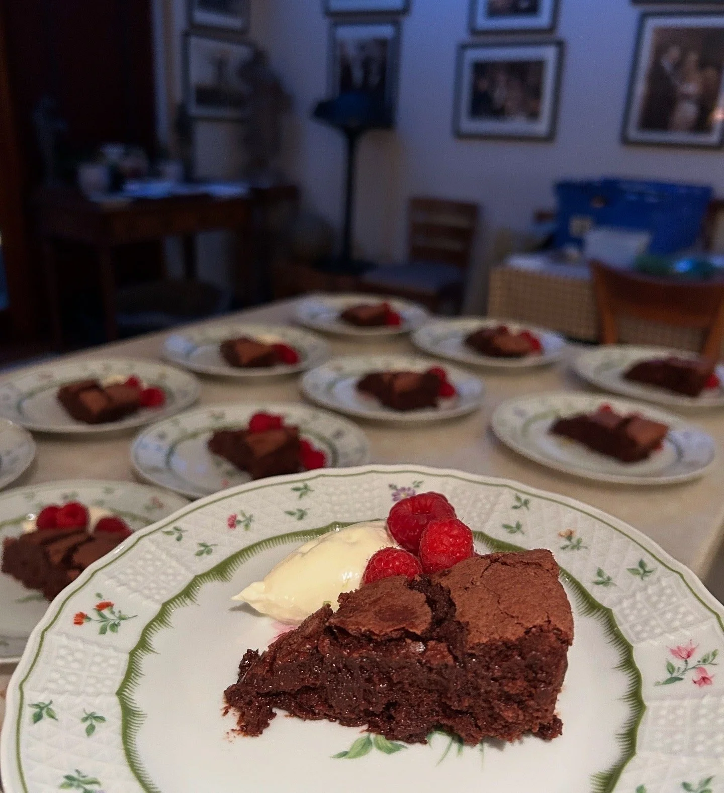 A slice of chocolate cake garnished with raspberries and a dollop of cream on a decorative plate, with several similar plates of cake in the background.