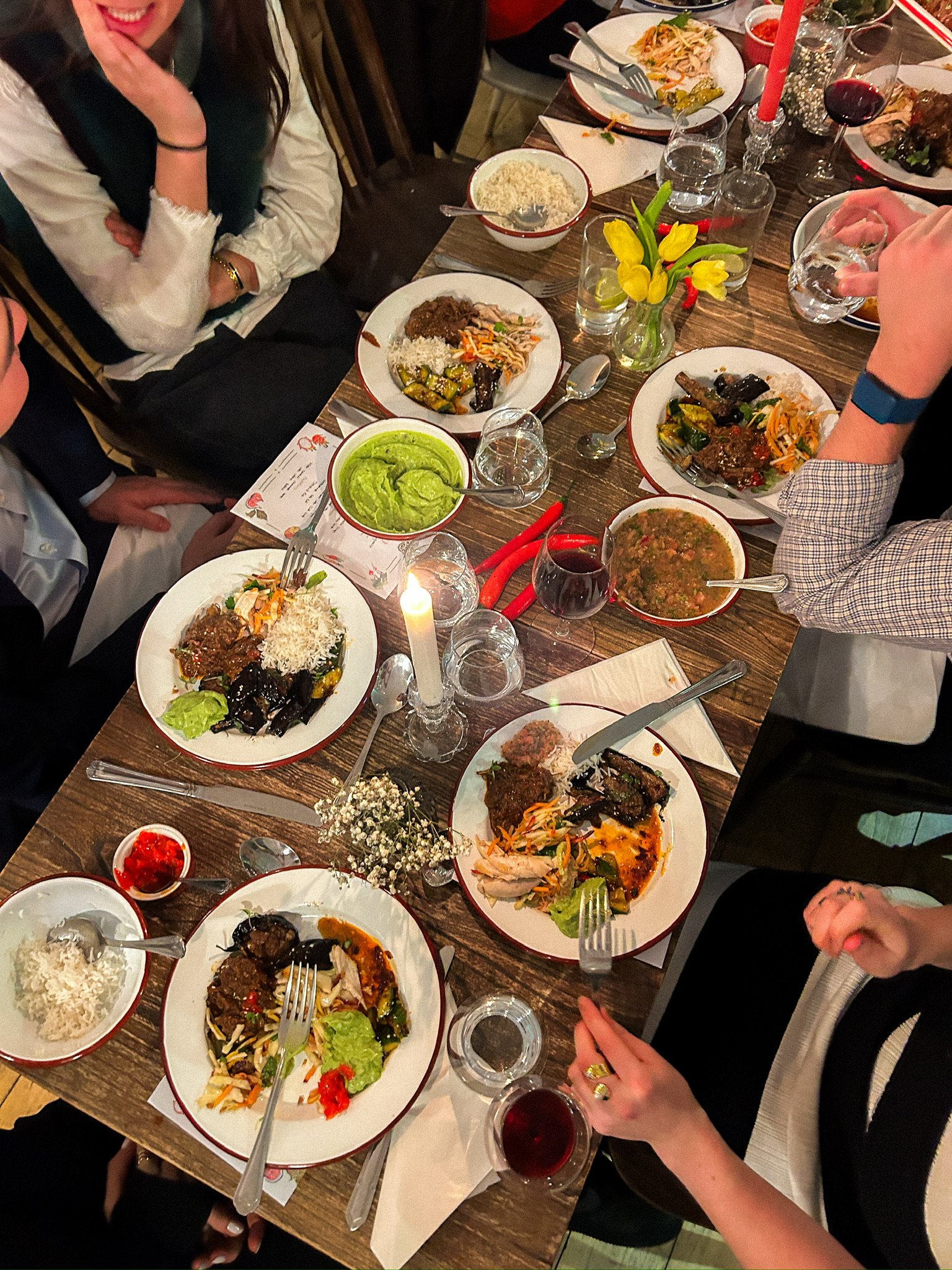 Dinner table with dishes of mixed vegetables, rice, guacamole, and sauces, surrounded by people with glasses of water and wine, candles and flowers on the table.
