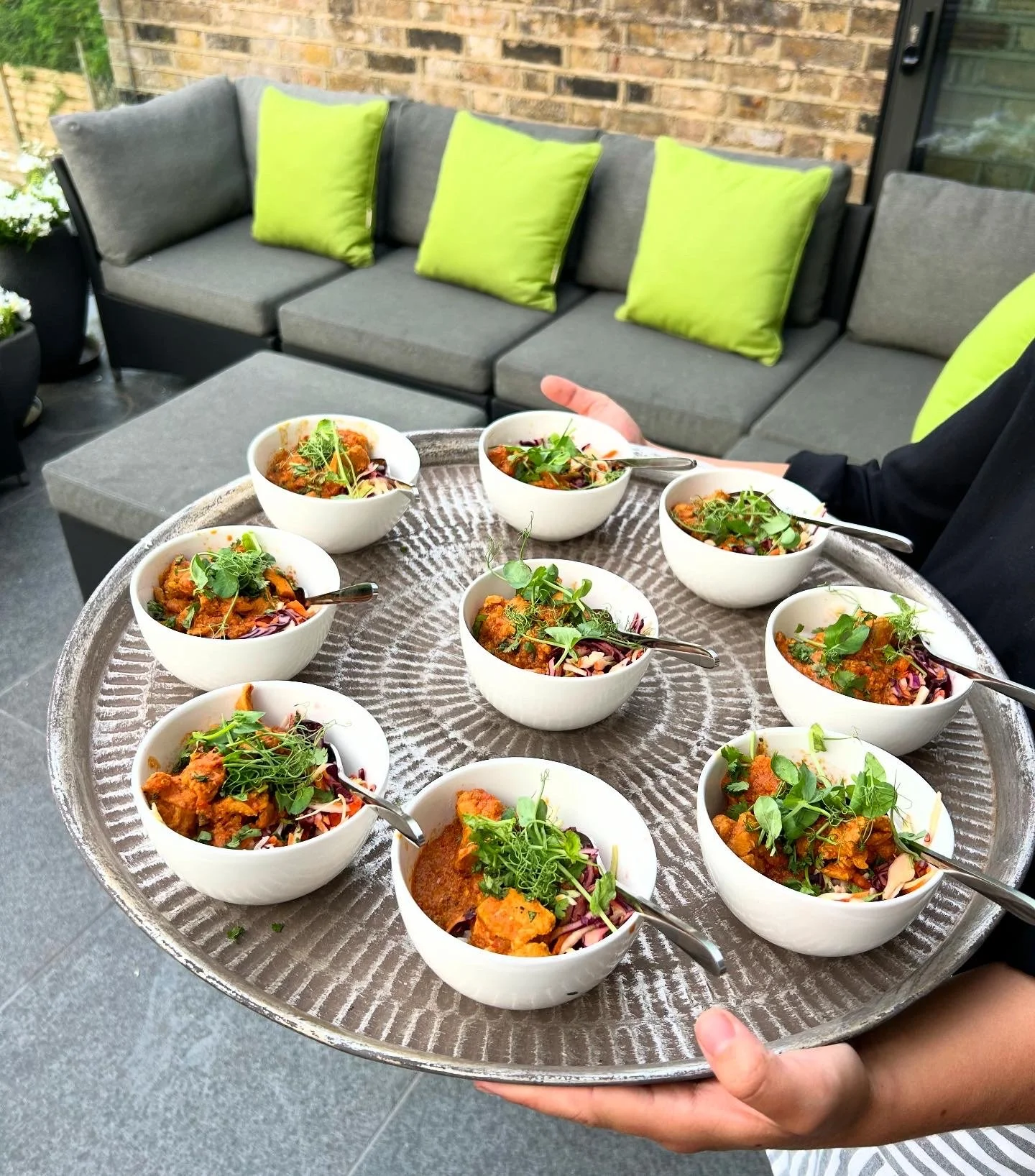 Person holding a large round tray with nine bowls of vegetable curry garnished with fresh greens, on a patio with a gray sectional sofa and green pillows, brick wall in the background.