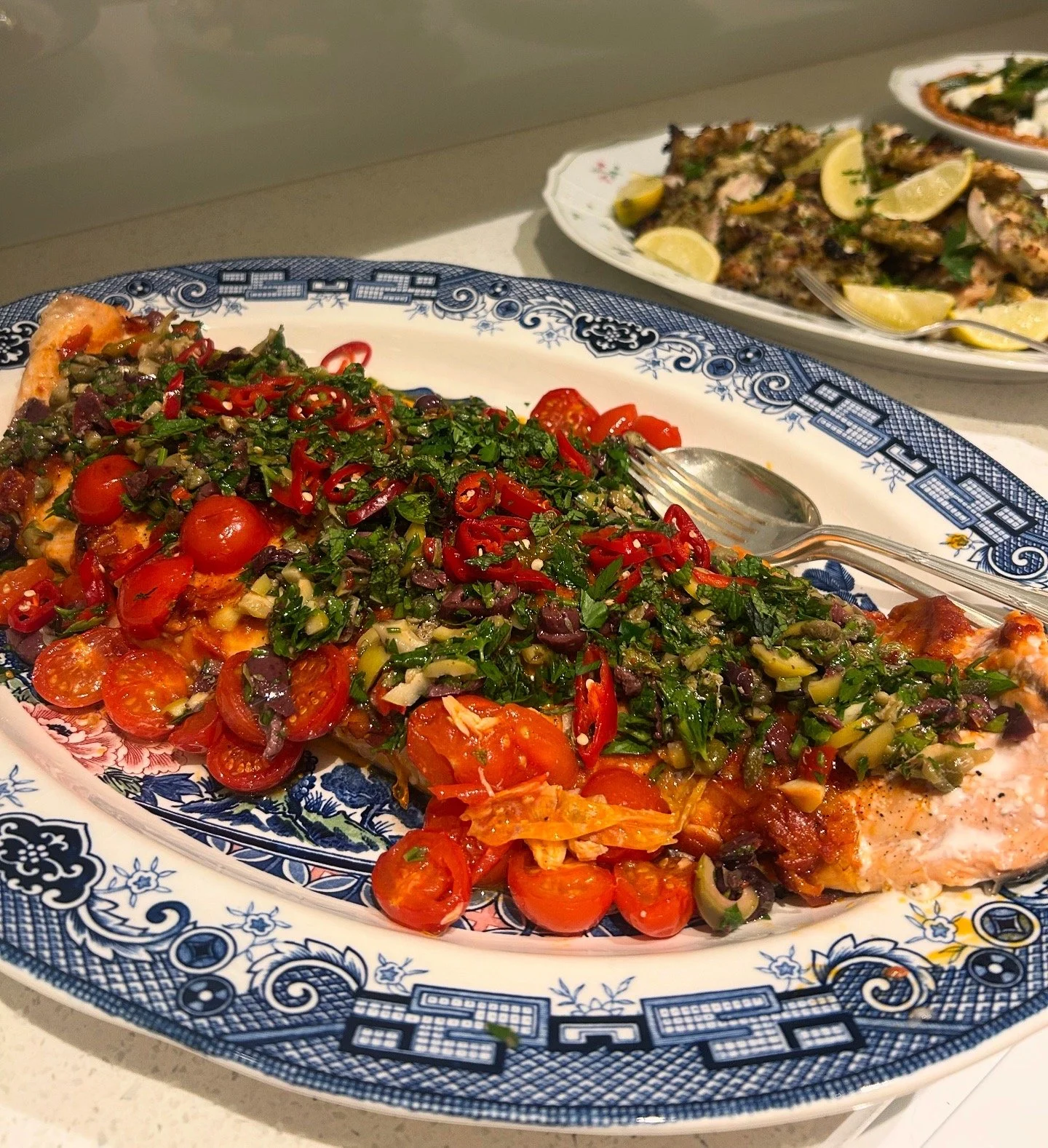 A large, whole baked fish covered with chopped fresh herbs, cherry tomatoes, and sliced red chili peppers, served on a decorative blue and white platter. In the background, there is another plate with lemon wedges and grilled fish.