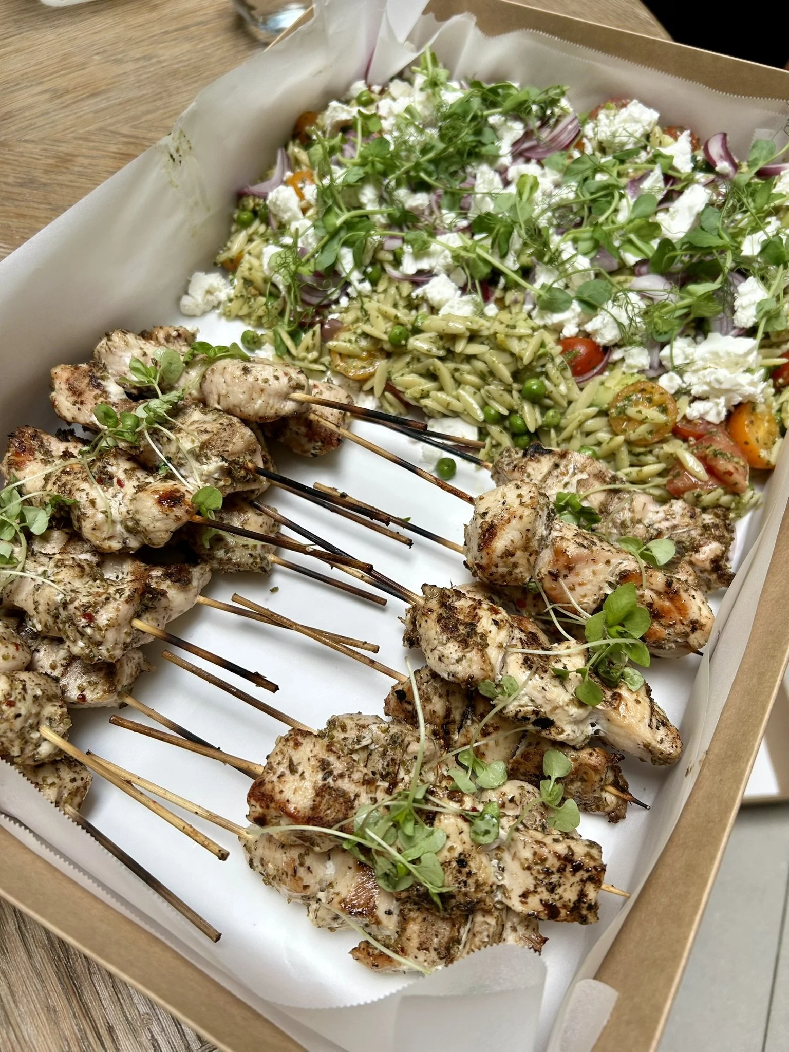 Grilled chicken skewers garnished with microgreens on a white tray with pasta salad containing cherry tomatoes, peas, feta cheese, and herbs.
