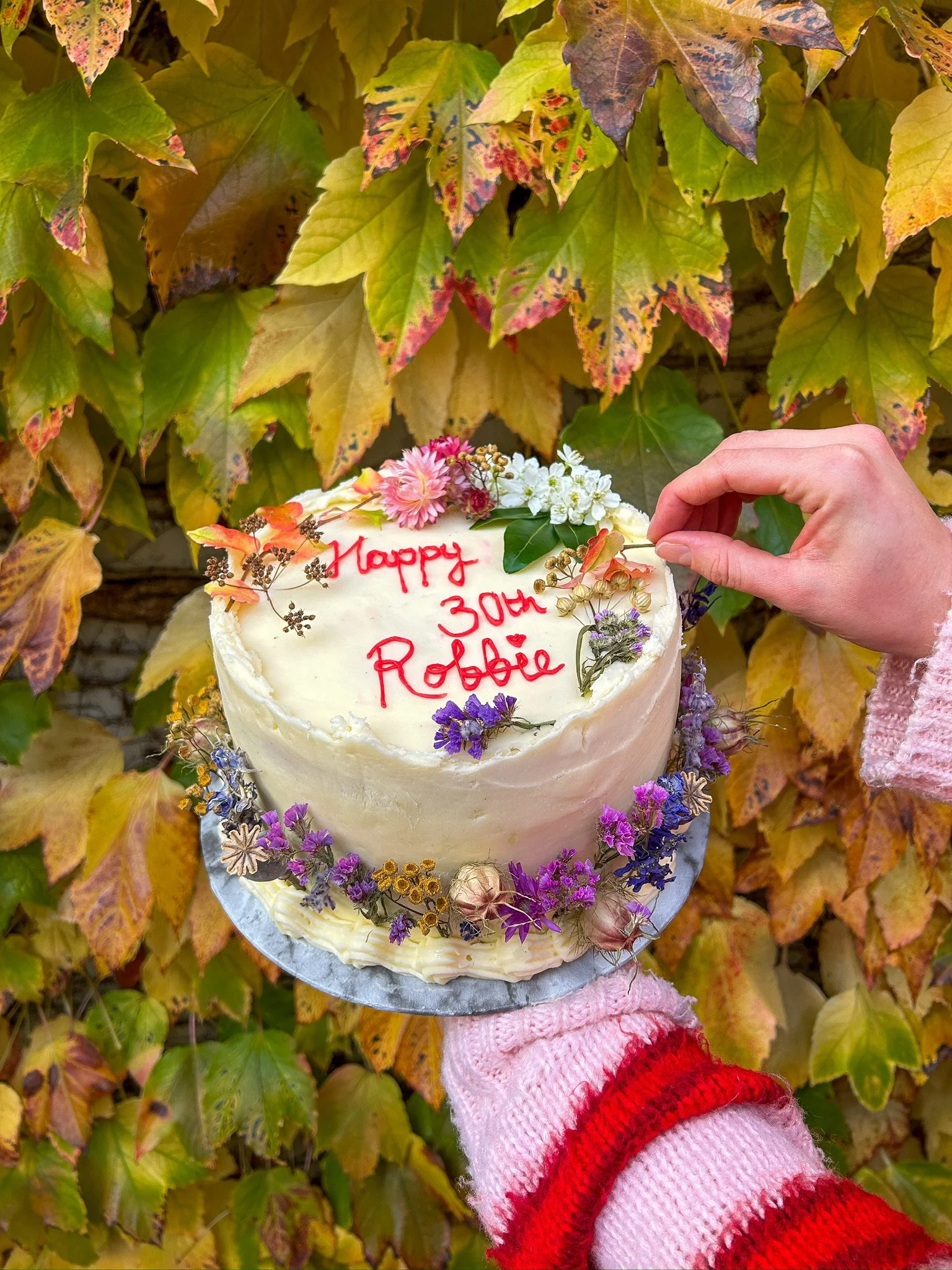 A birthday cake with white frosting decorated with flowers, being held in front of colorful autumn leaves. The cake has red writing that reads 'Happy 30th Robbìe'. A person wearing pink and red striped sweater is placing a flower on the cake.